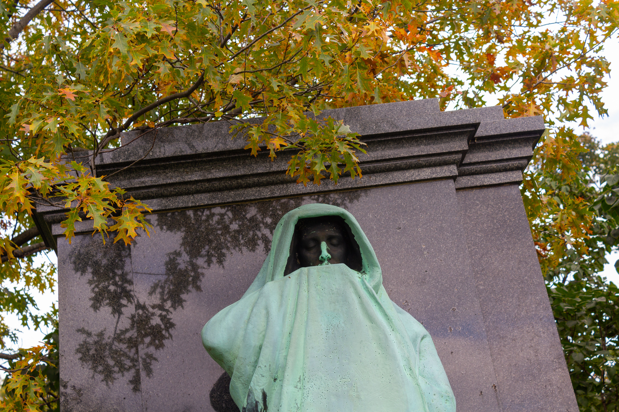 Head and shoulders of a cloaked bronze statue, green with age, against the top of a black tomb and yellow tree leaves
