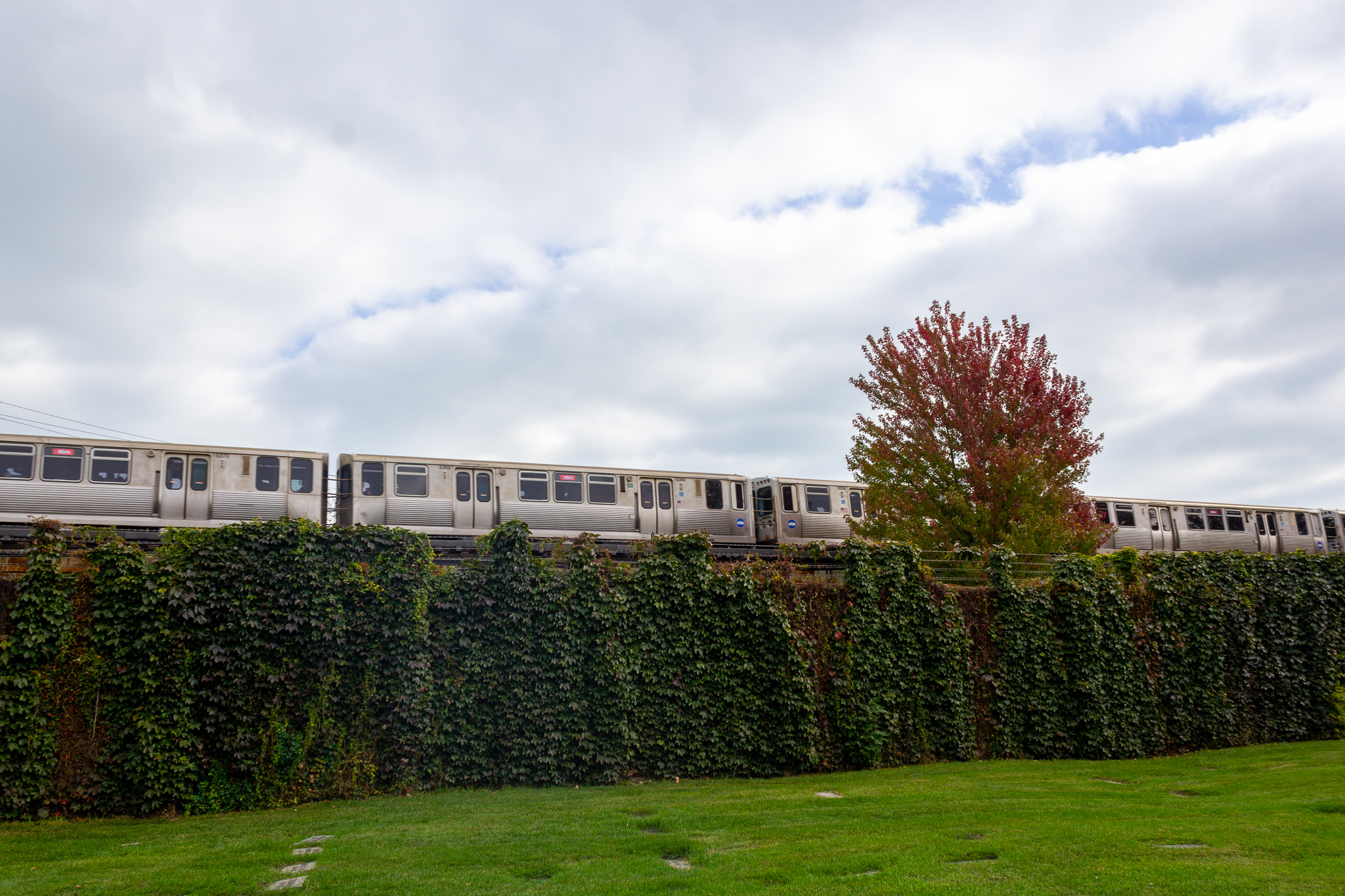 An elevated train over an ivy-covered wall, green cemetery grounds in the foreground and a clouded pale blue sky in the background