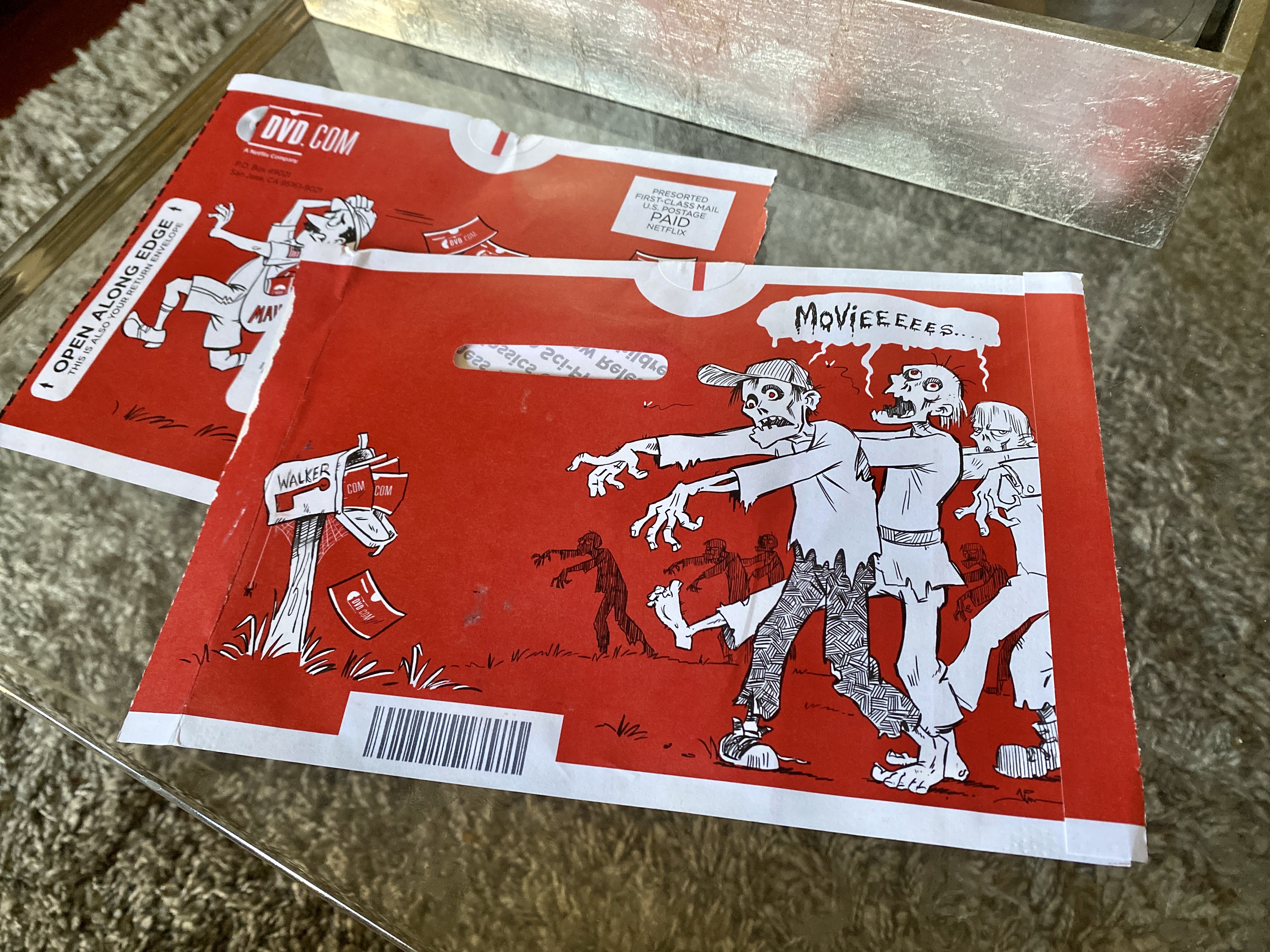 Red DVD.com mailing sleeve with a black and white cartoon illustration of zombies lumbering towards a mailbox filled with DVDs; a speech bubble above their heads says Movieeeees