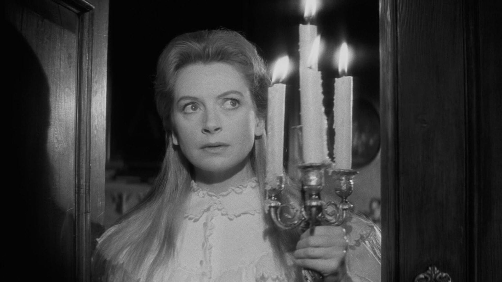Still from film THE INNOCENTS with a white woman with long blond hair and an old-fashioned nightgown, holding a large candelabra, peering apprehensively beyond the camera