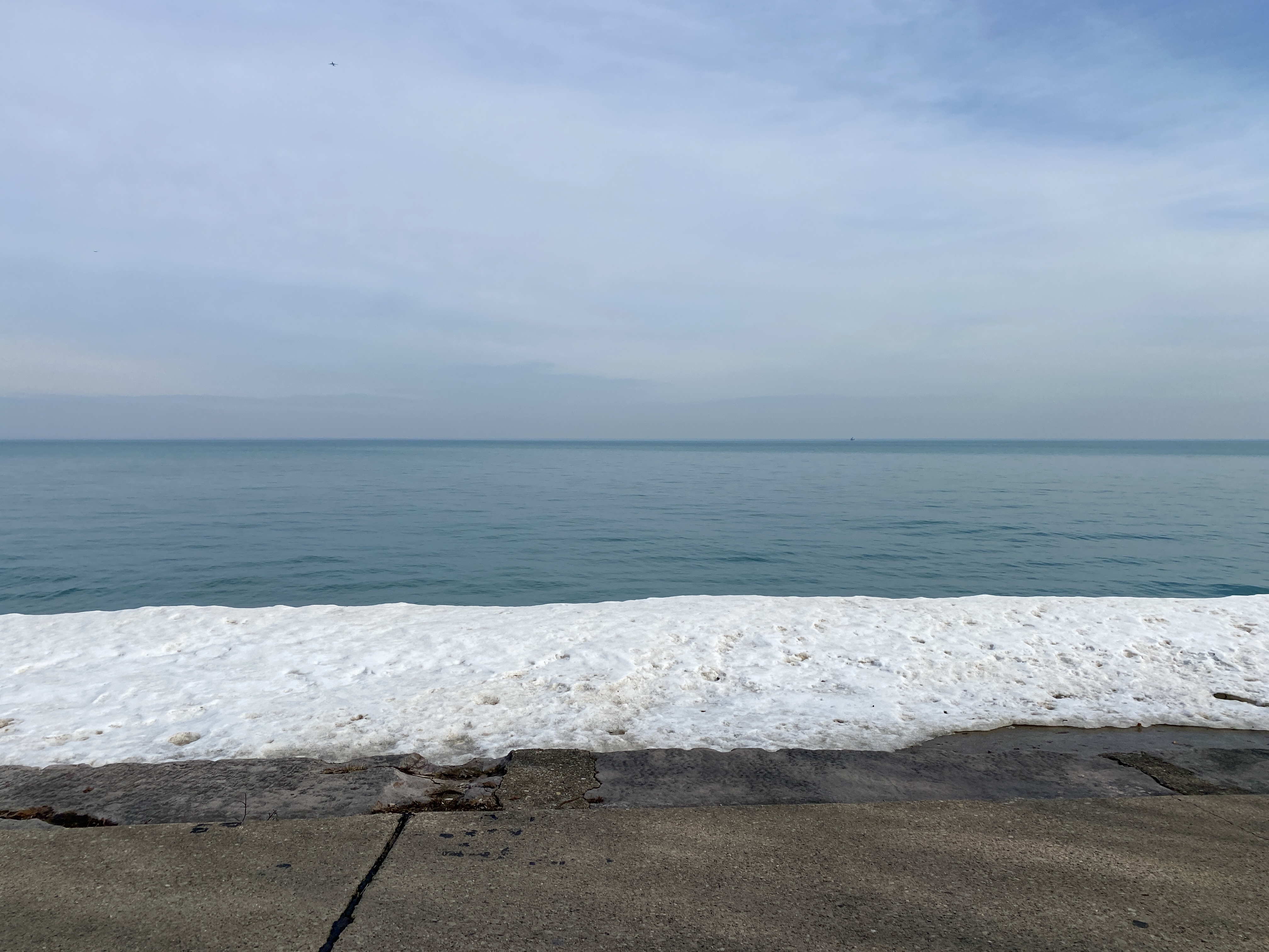 Horizon of large Lake Michigan with cloud-swept pale blue sky and a shelf of snow in the foreground