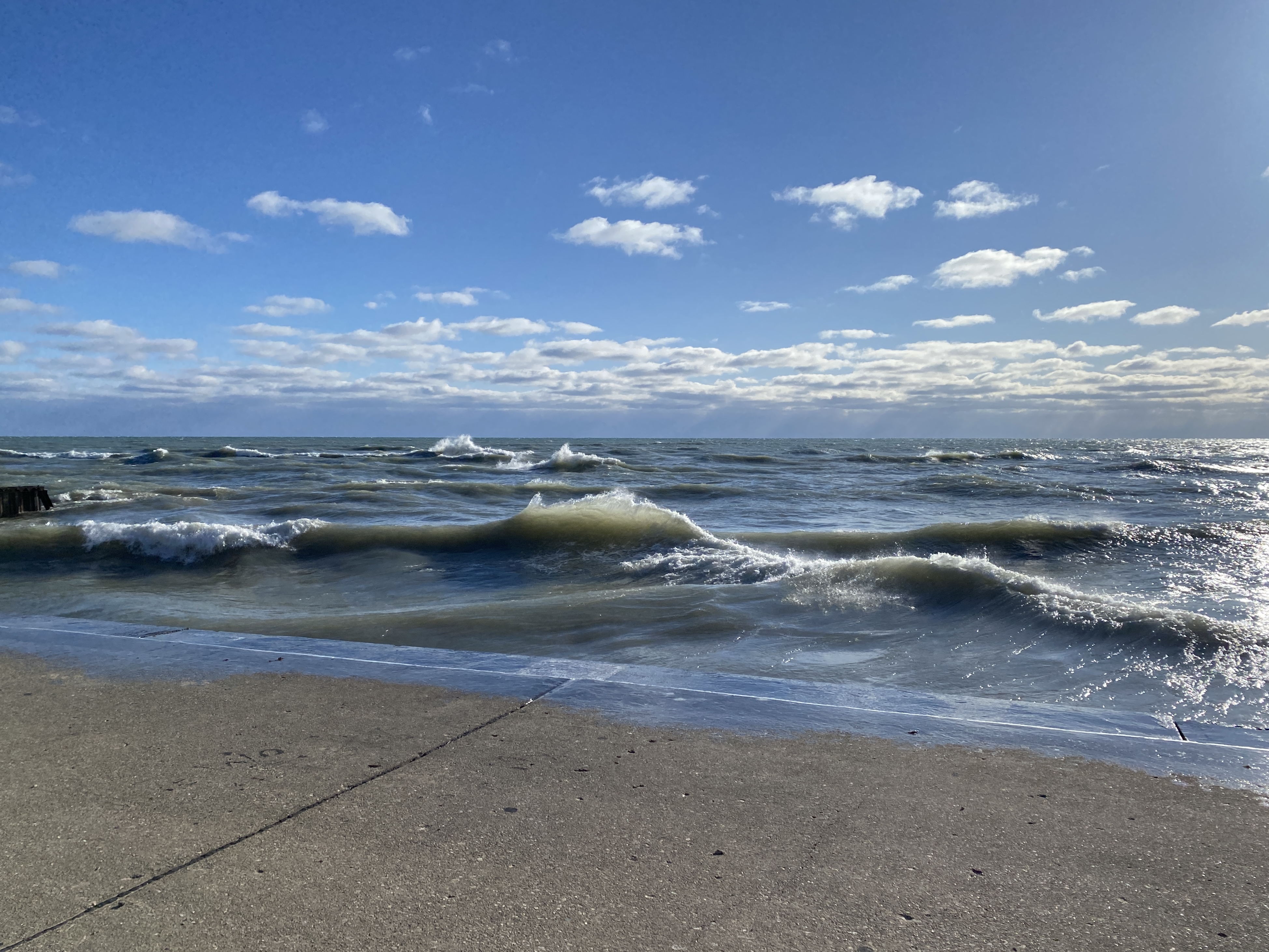 Angled horizon of large Lake Michigan with a blue sky dotted with small white clouds overhead, pronounced waves and a concrete shelf in the foreground