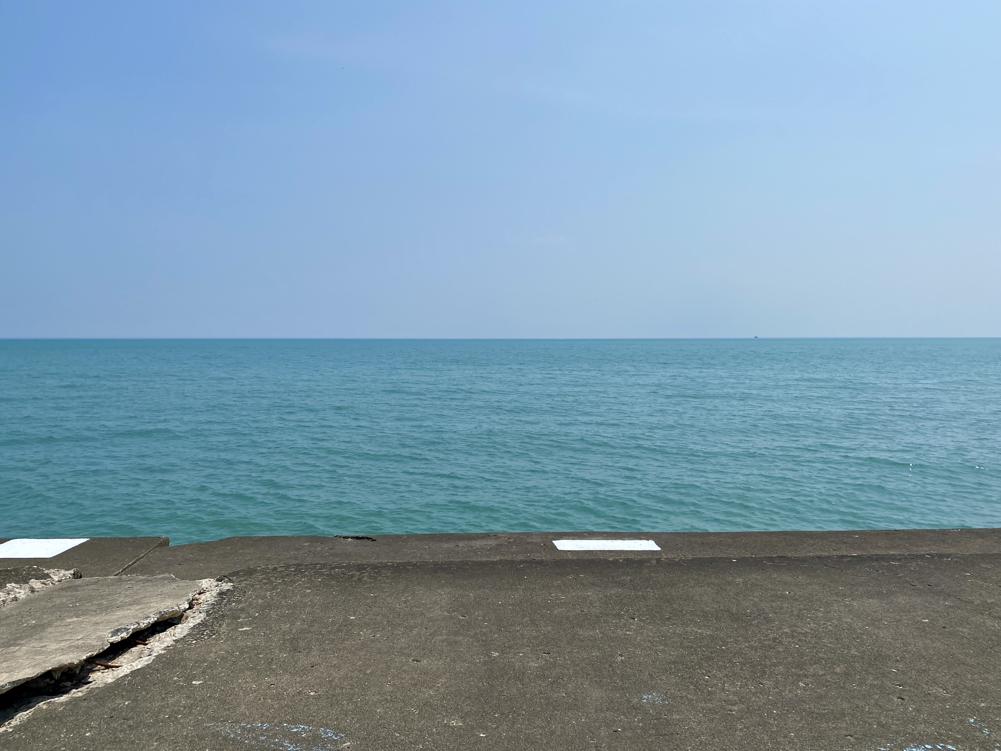 Horizon of Lake Michigan with a hazy blue sky above calm darker blue water and an concrete shelf in the foreground