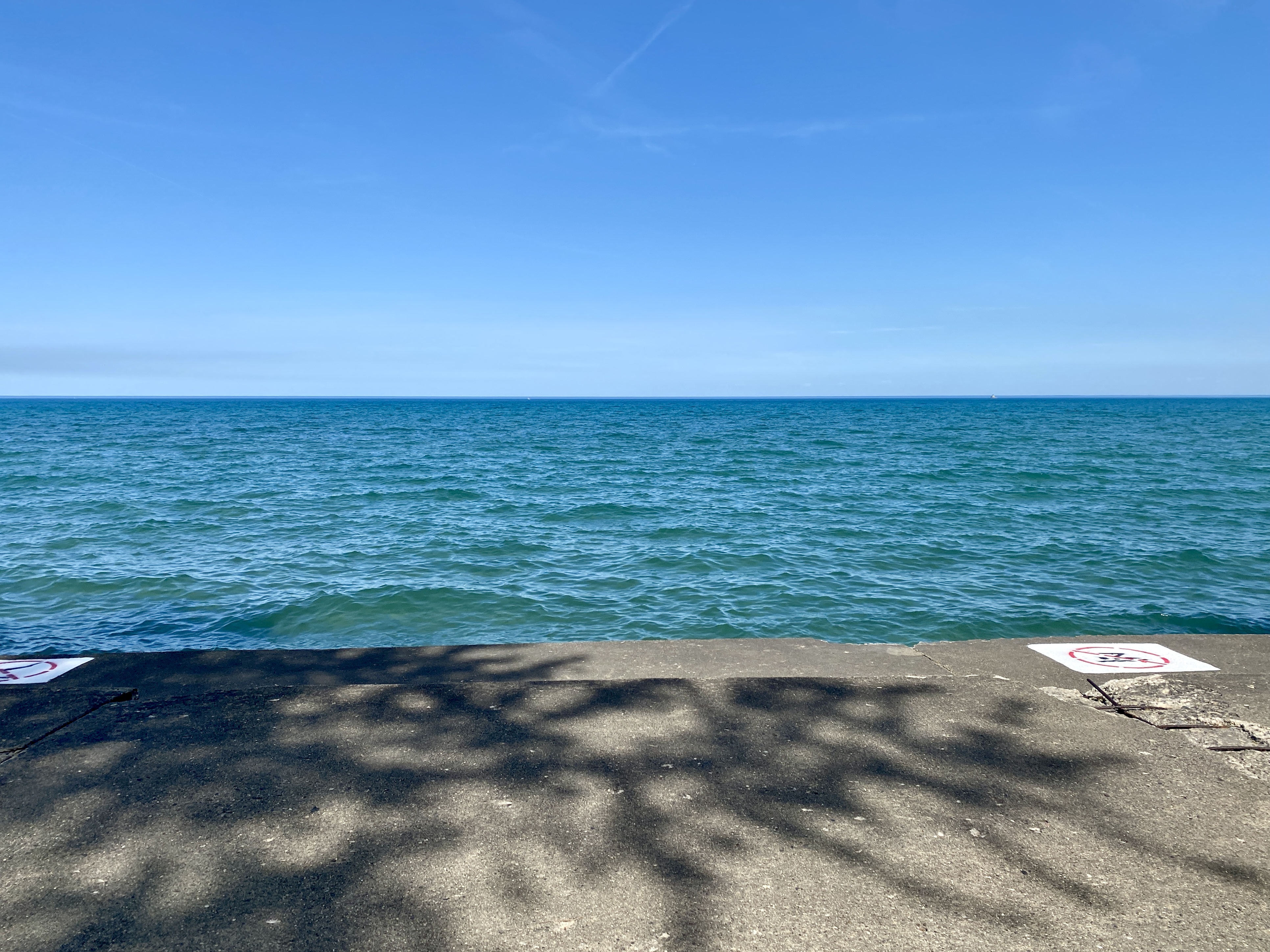 Horizon of large Lake Michigan with a deep, clear blue sky and turquoise-shaded water with small waves, a grey concrete shelf in the foreground