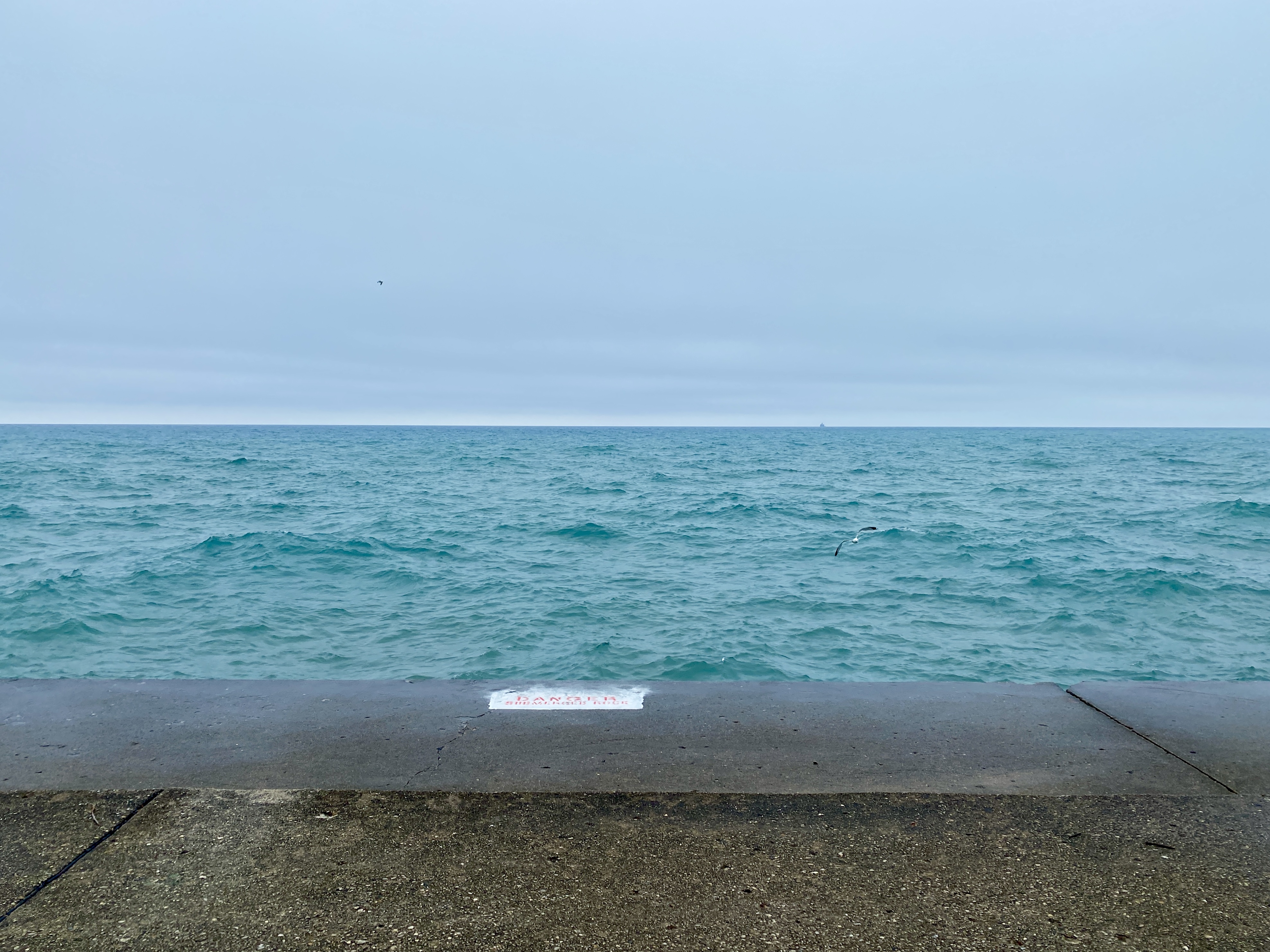 Horizon of a cloudy pale blue sky meeting a large lake with moderate waves of turquoise with a strip of concrete in the foreground