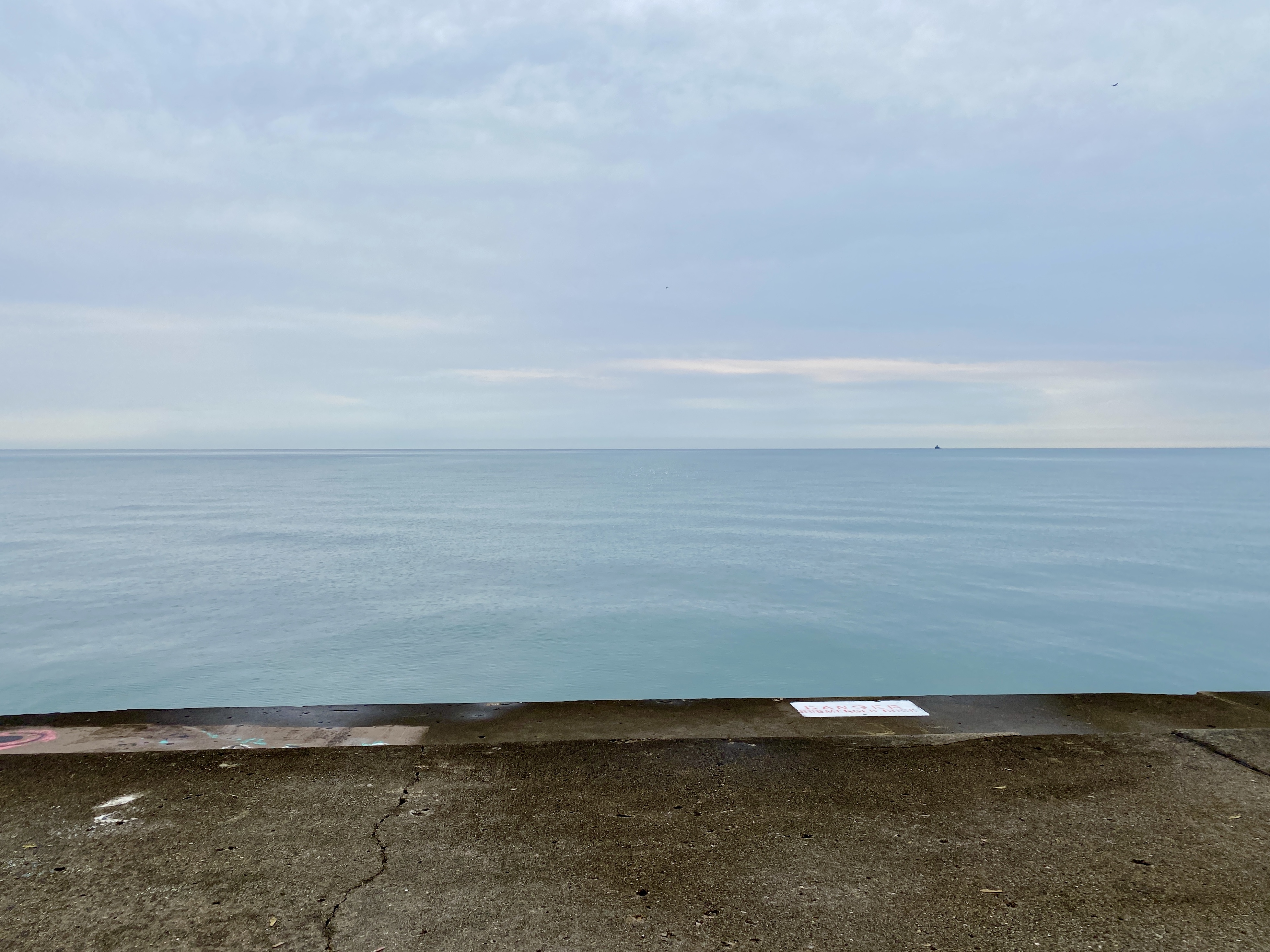 Horizon of a cloudy pale blue sky meeting a large lake with a smooth blue surface, slight pink from a sunrise glinting through, and a strip of concrete in the foreground