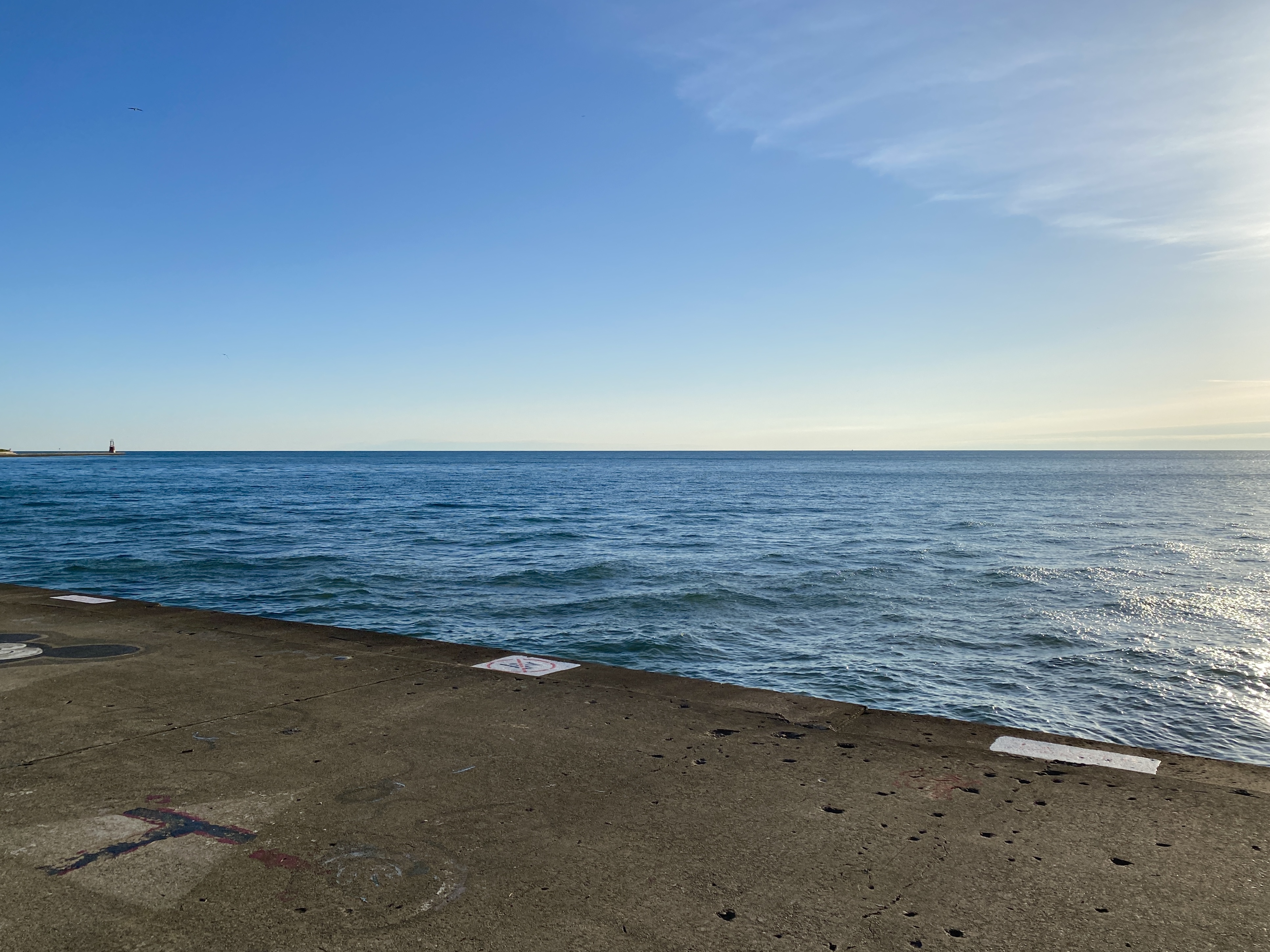 Horizon of a blue sky meeting a large, deep blue lake with small waves, pale yellow light from an early morning sun shining to the left and a strip of concrete angled in the foreground
