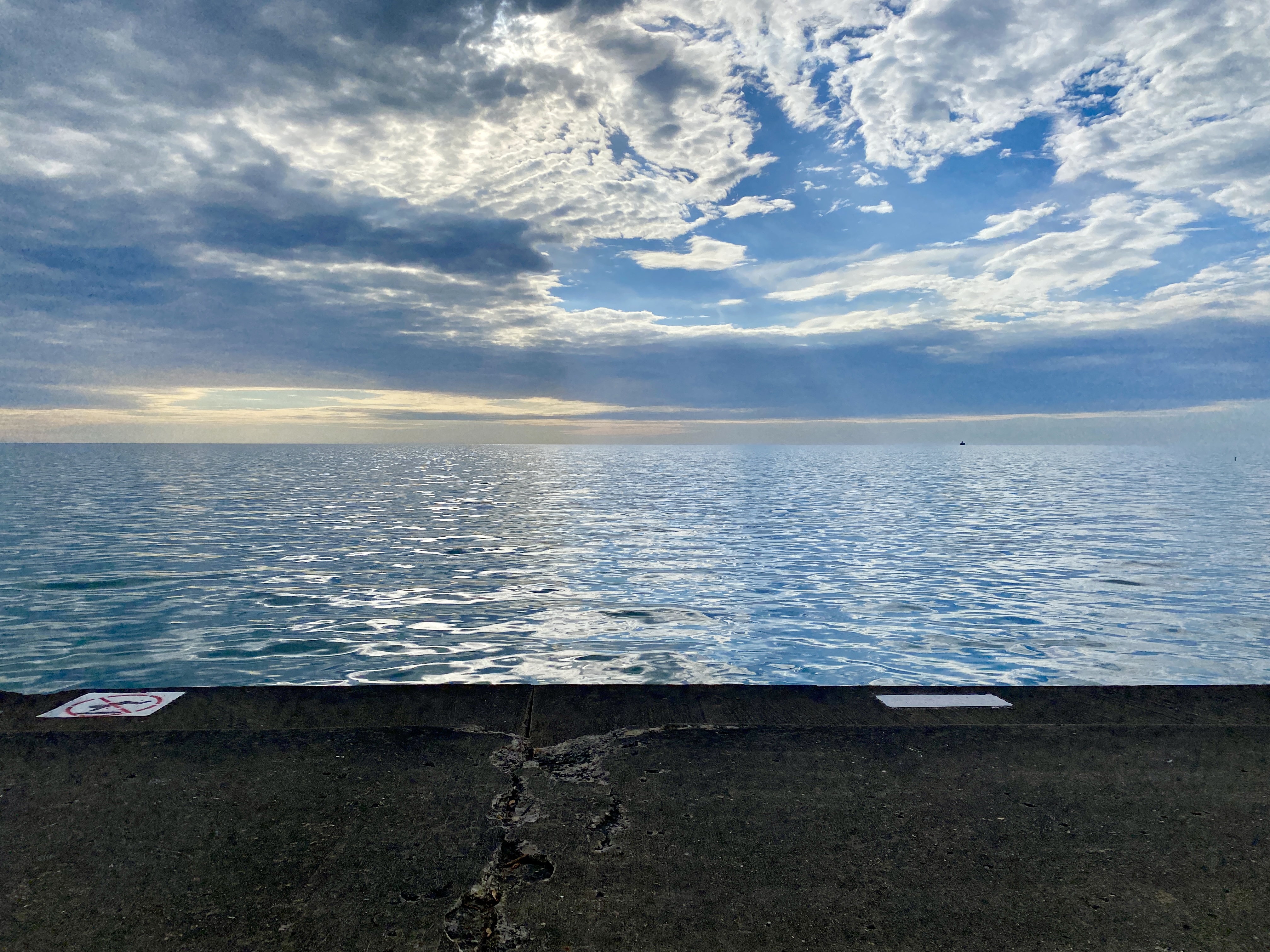 Horizon of a large lake with clouds swept over a blue sky and a strip of yellow light with the early morning sun, water smooth, and a strip of concrete in the foreground
