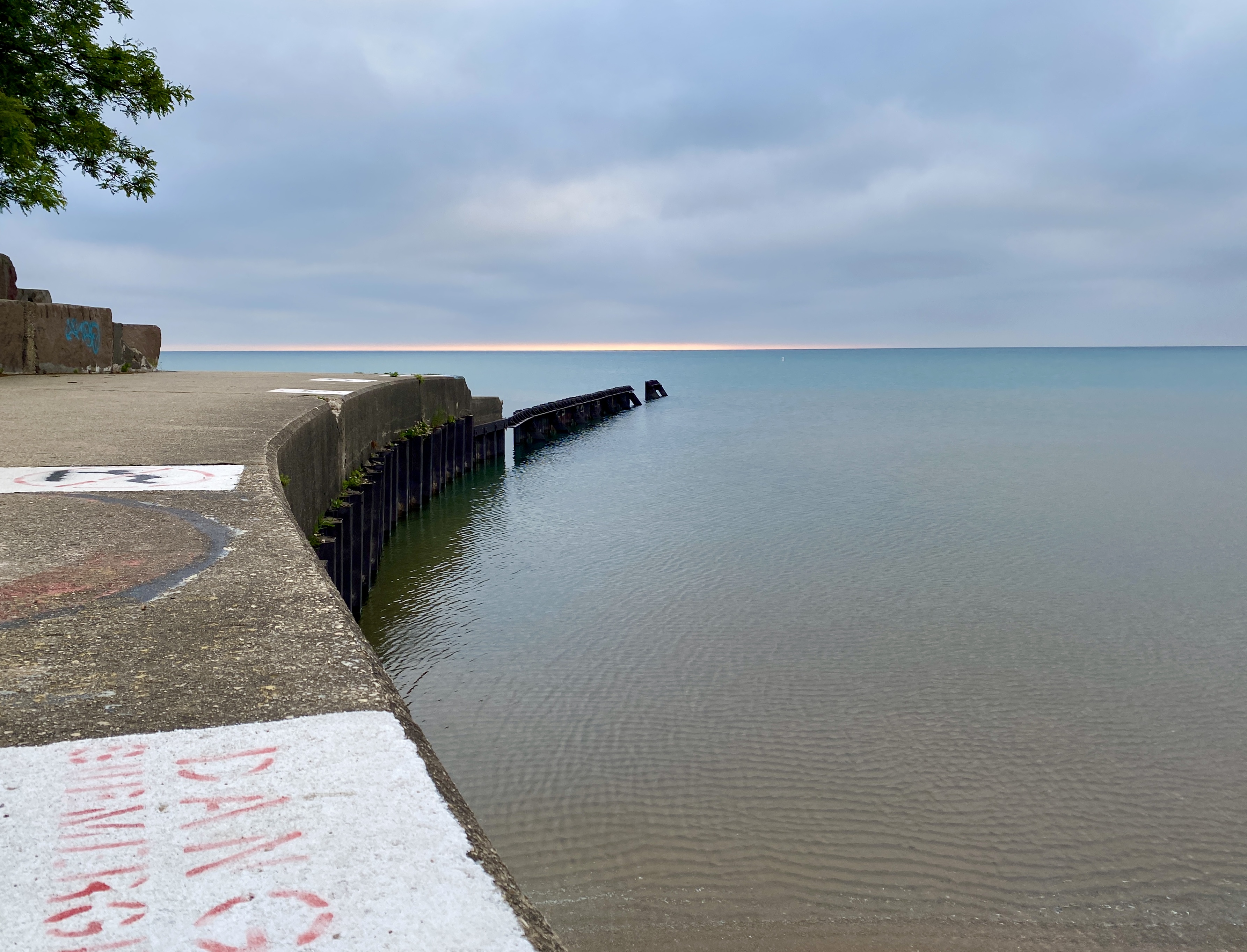 Horizon of a large lake with a clouded sky, a thin strip of orange sunrise, and a curve of concrete walkway to the left with a green tree, the water still blue fading to sandy brown at the beach on the right