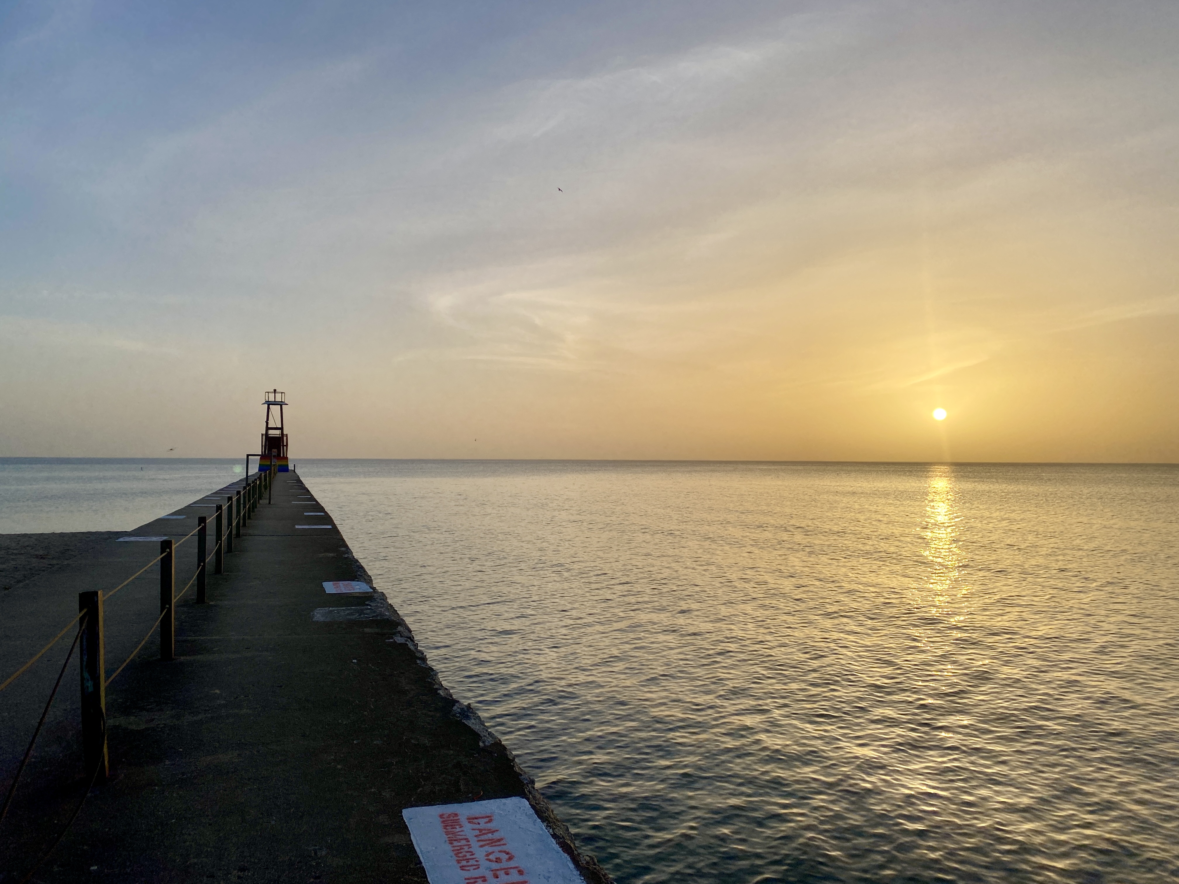 Horizon of a large lake, a narrow concrete pier jutting out to the left and a sun rising to the right, the hazy sky and water tinted warm yellow