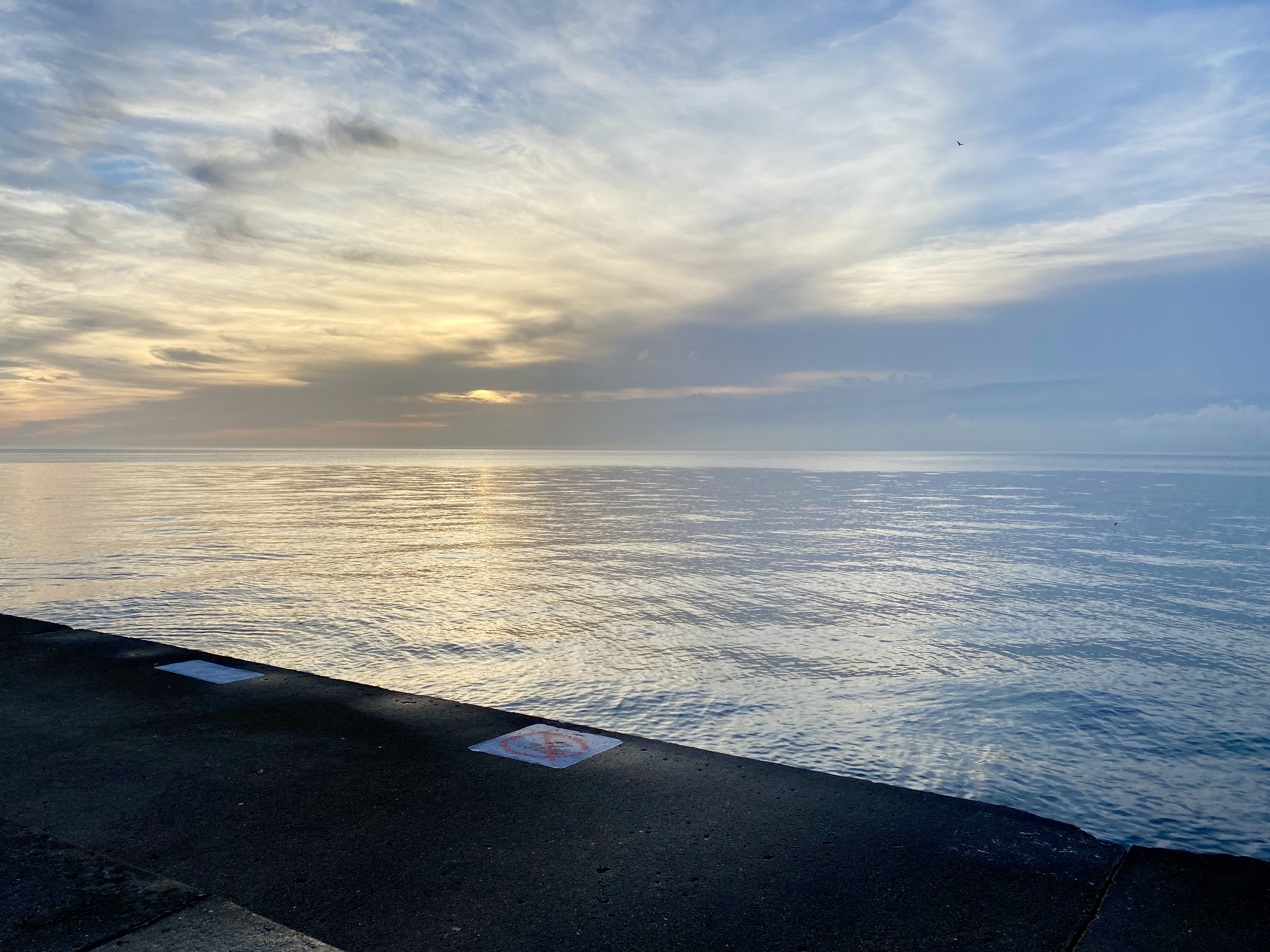 Horizon of a large blue lake below a sky swept with clouds, a pale yellow sunrise peeking through, and an angled strip of dark concrete walkway