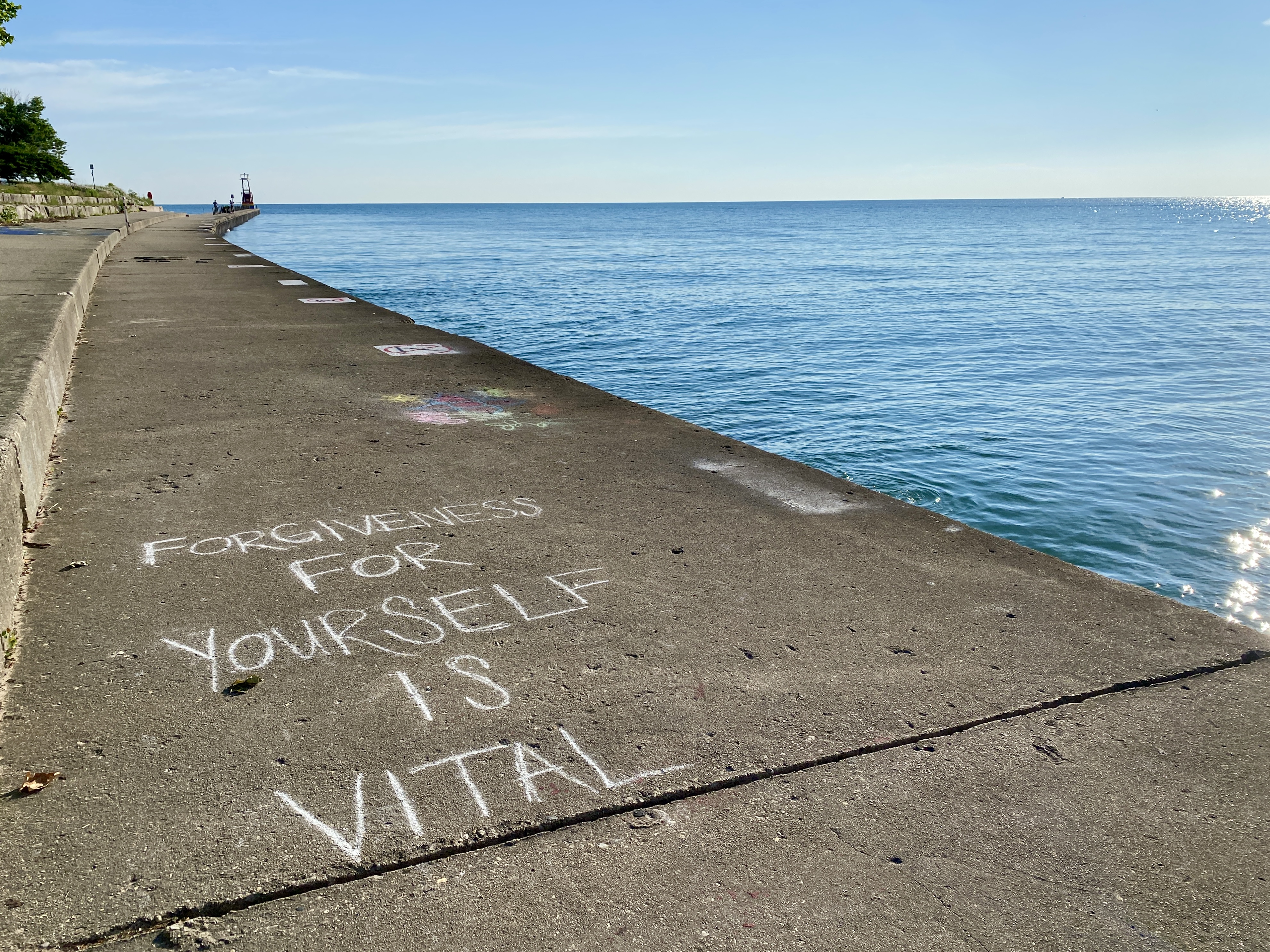 Lakeshore, lined with concrete, curving to the left, to right bright blue lake surface below a blue sky; written on the concrete in the foreground is FORGIVENESS FOR YOURSELF IS VITAL