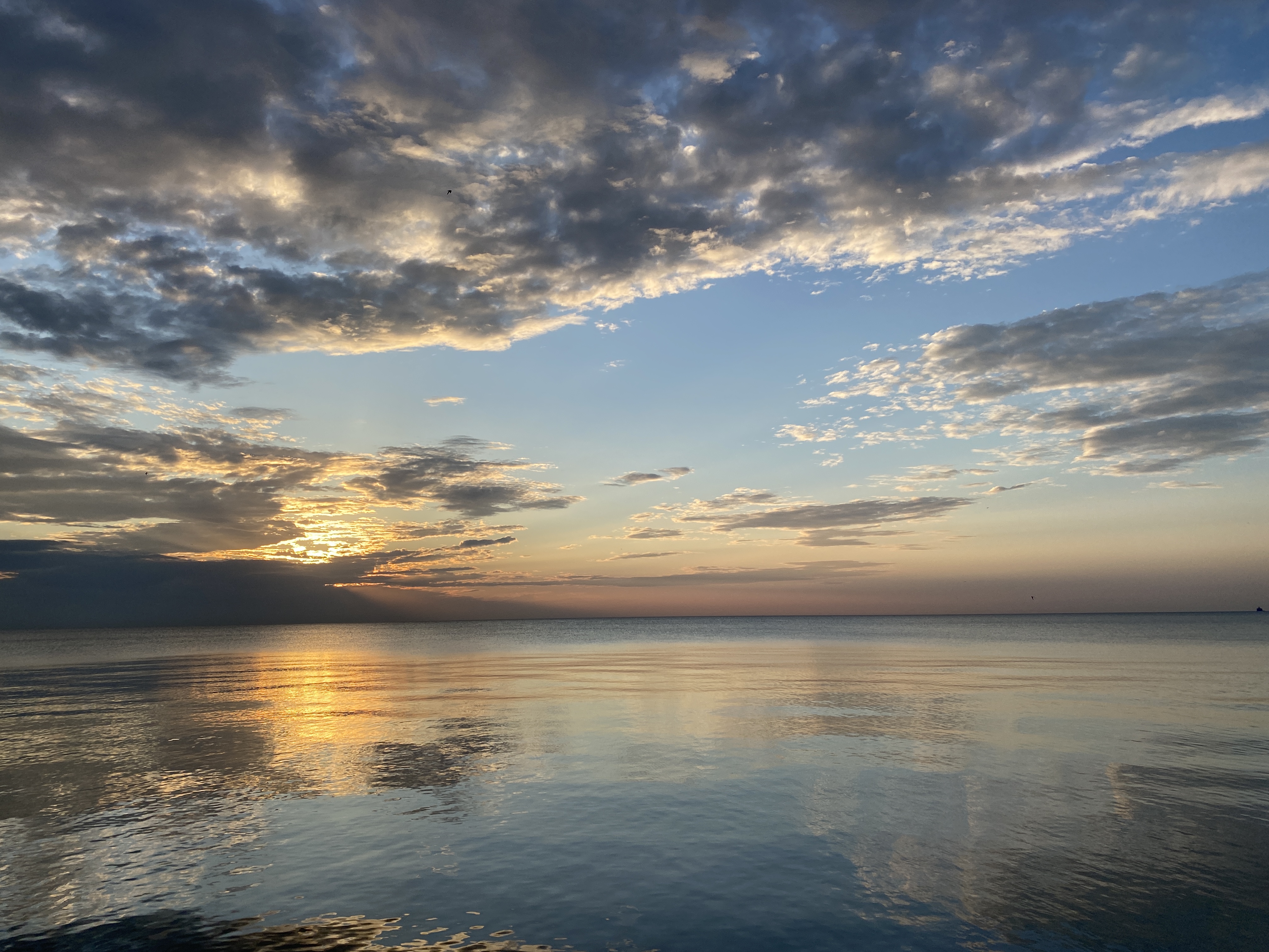 A large lake under a blue sky swept with broken clouds, an orange sunrise just behind, the water surface smooth and glassy, reflecting the colors of the sky