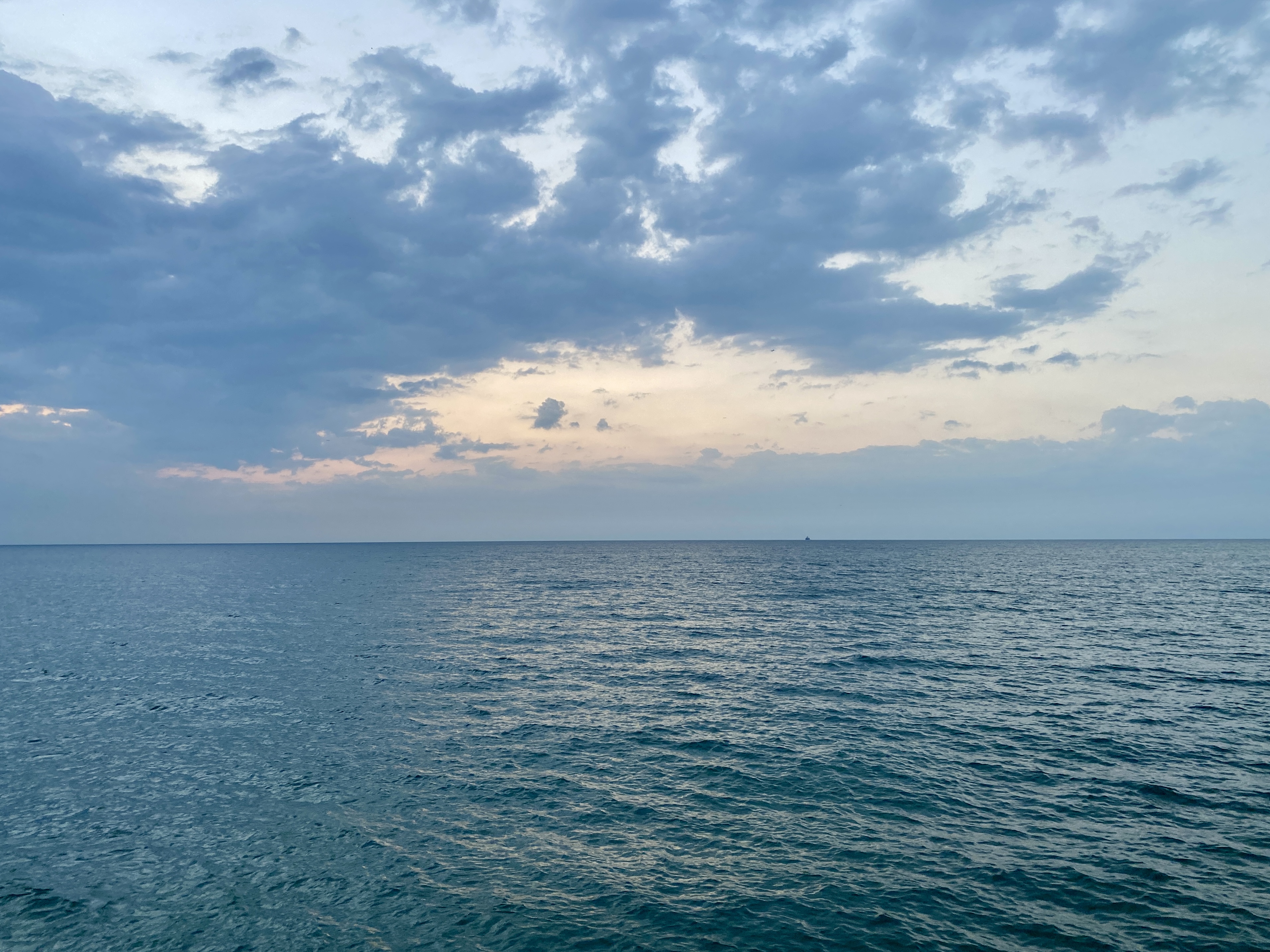 A large lake under a soft blue sky spotted with clouds, the water dark blue and calm, a hint of pink sunrise over the horizon