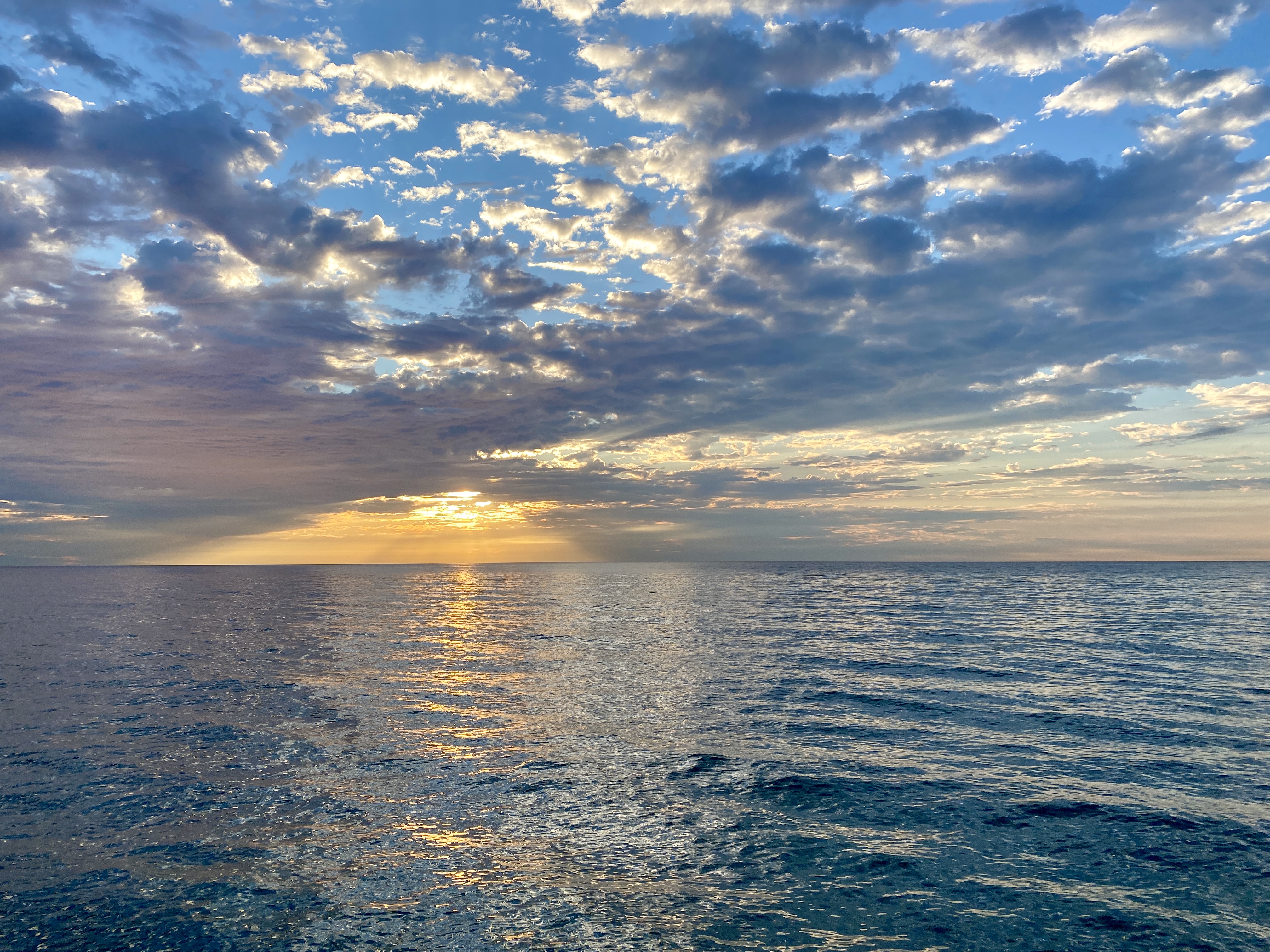 A lake horizon with an orange sunrise peeking through a blue sky swept with clouds, the water surface dark blue and calm