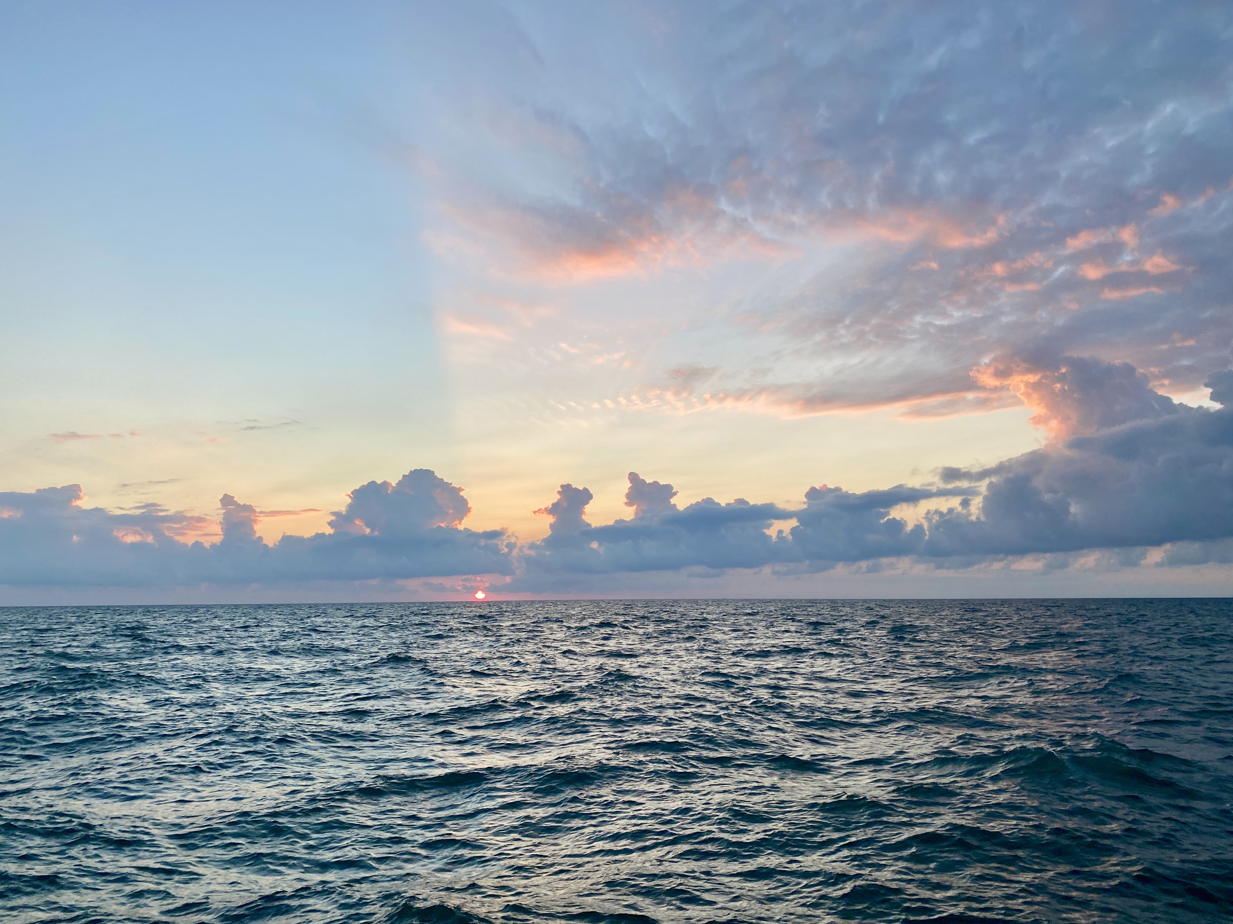 A round red sun rising over a lake horizon, the sky pale blue and swept with yellow and rose clouds, the water surface dark blue with moderate waves