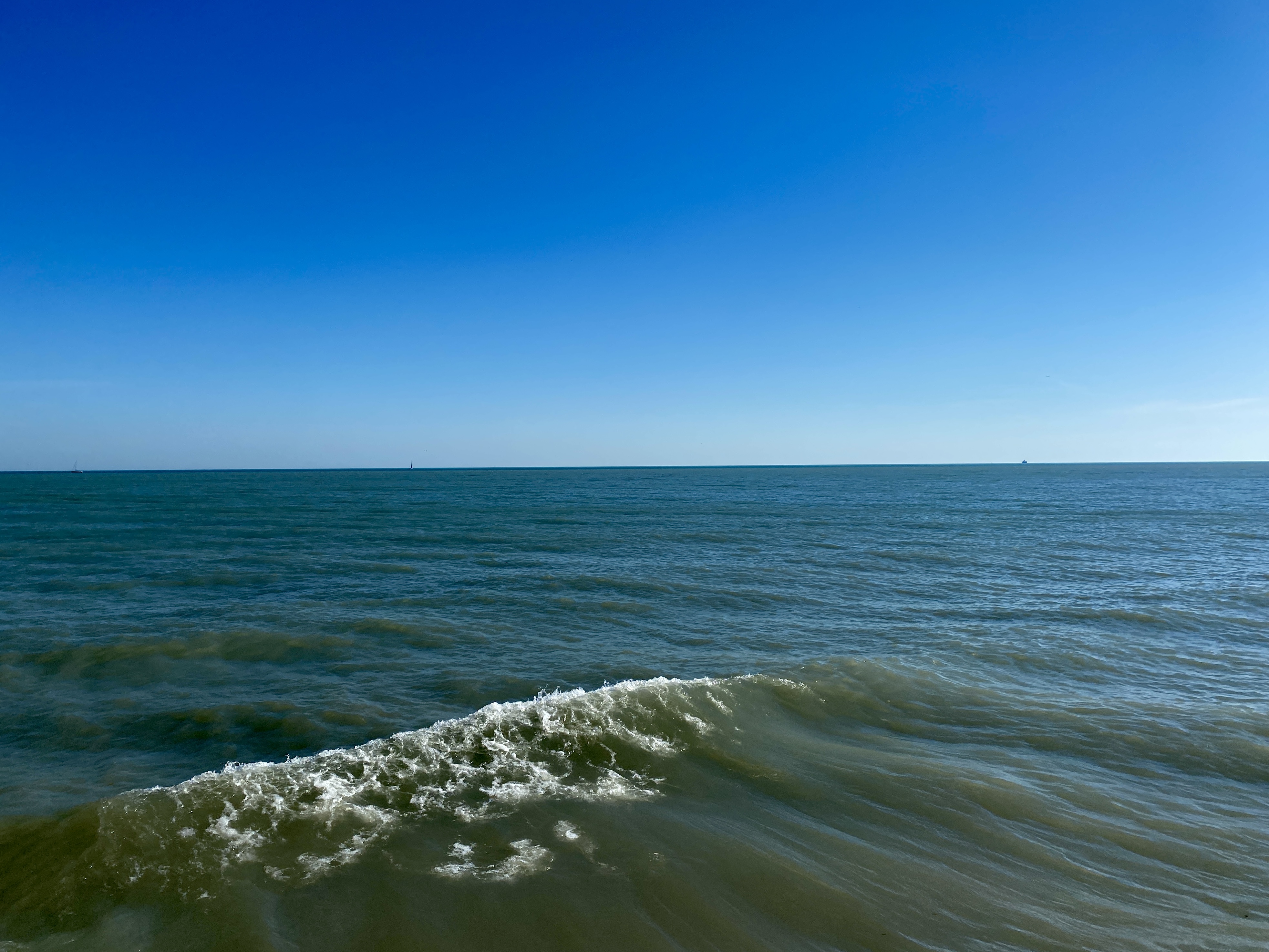 A wide lake horizon with a clear, bright blue sky and a slanted line of a wave in the deep green water