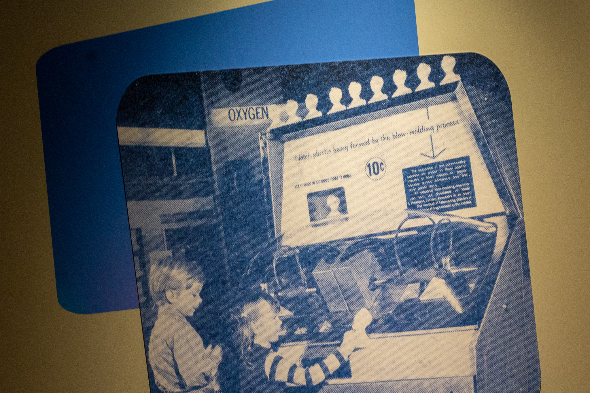A large photo of a children using a Mold-A-Rama machine in the 1960s mounted on a grey wall