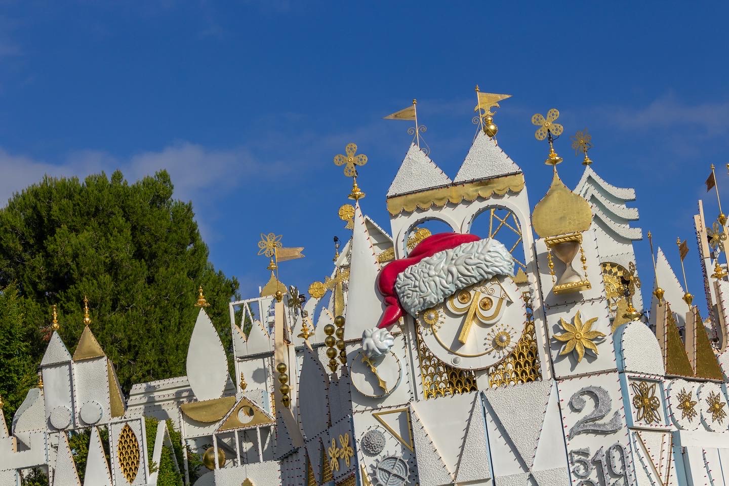 White textured geometric shapes form the top edge of the It's a Small World ride against a blue sky and line of green trees; the round white and gold face in the middle wears a large Santa hat