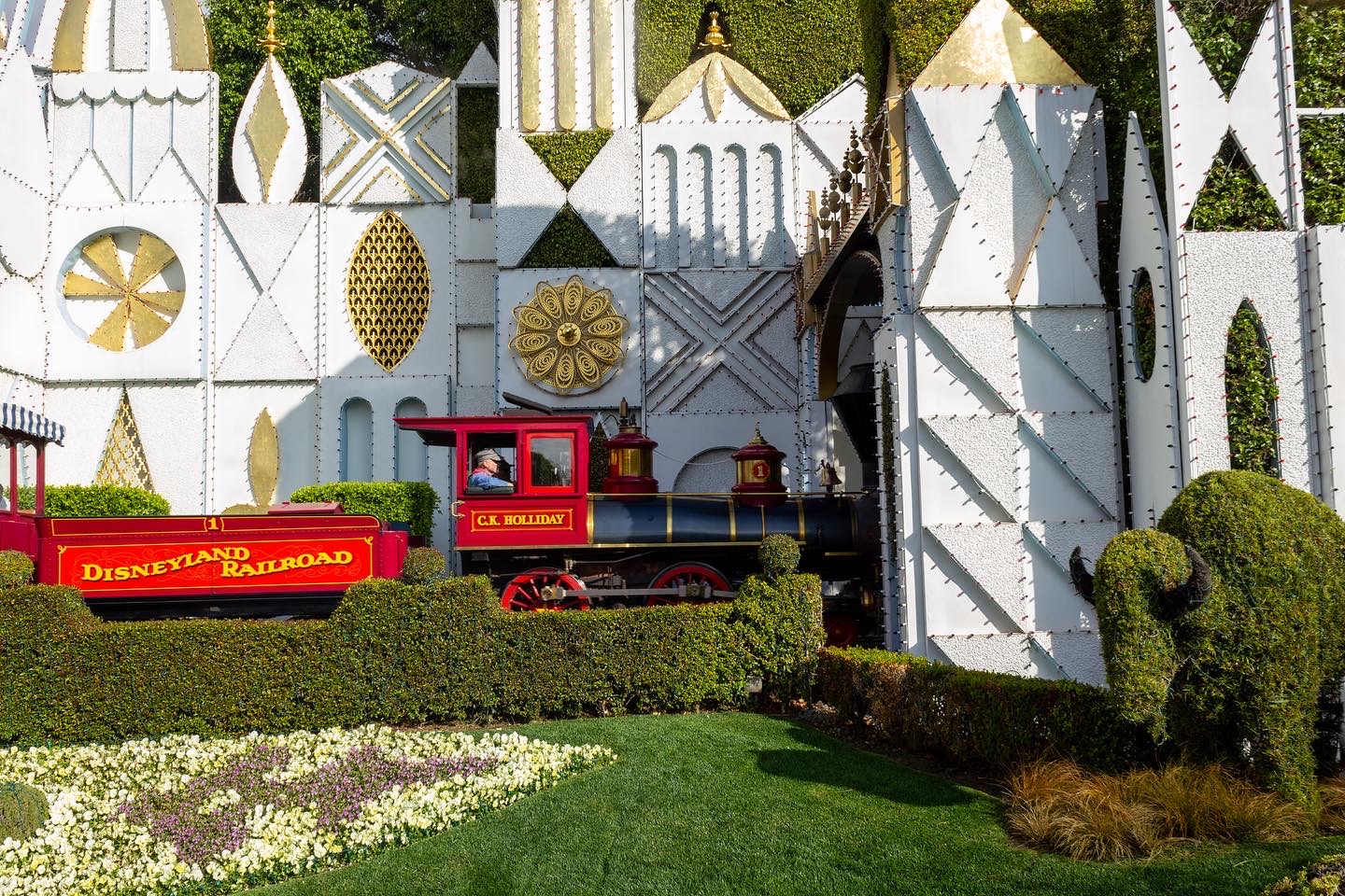 The bright red Disneyland Railroad train travels in front of the textured white and gold panels of the It's a Small World ride