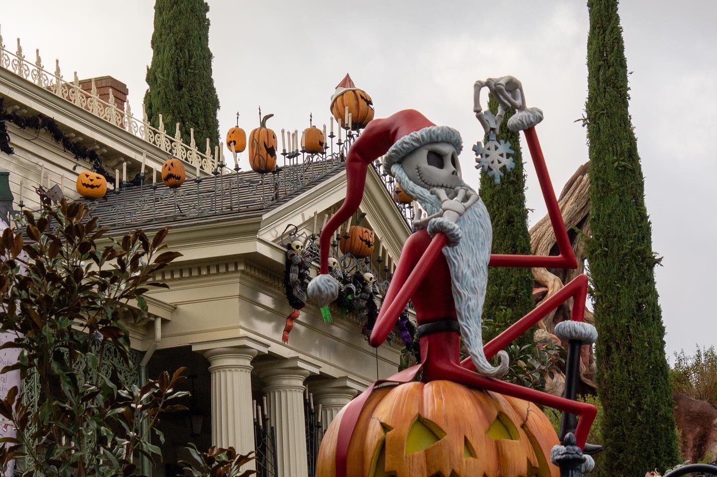 The thin skeleton figure of Jack Skellington, dressed in a red Santa outfit, perches on top of the gate to the Haunted Mansion; behind him, the white New Orleans-style mansion is decorated with black ribbons and orange jack o' lanterns