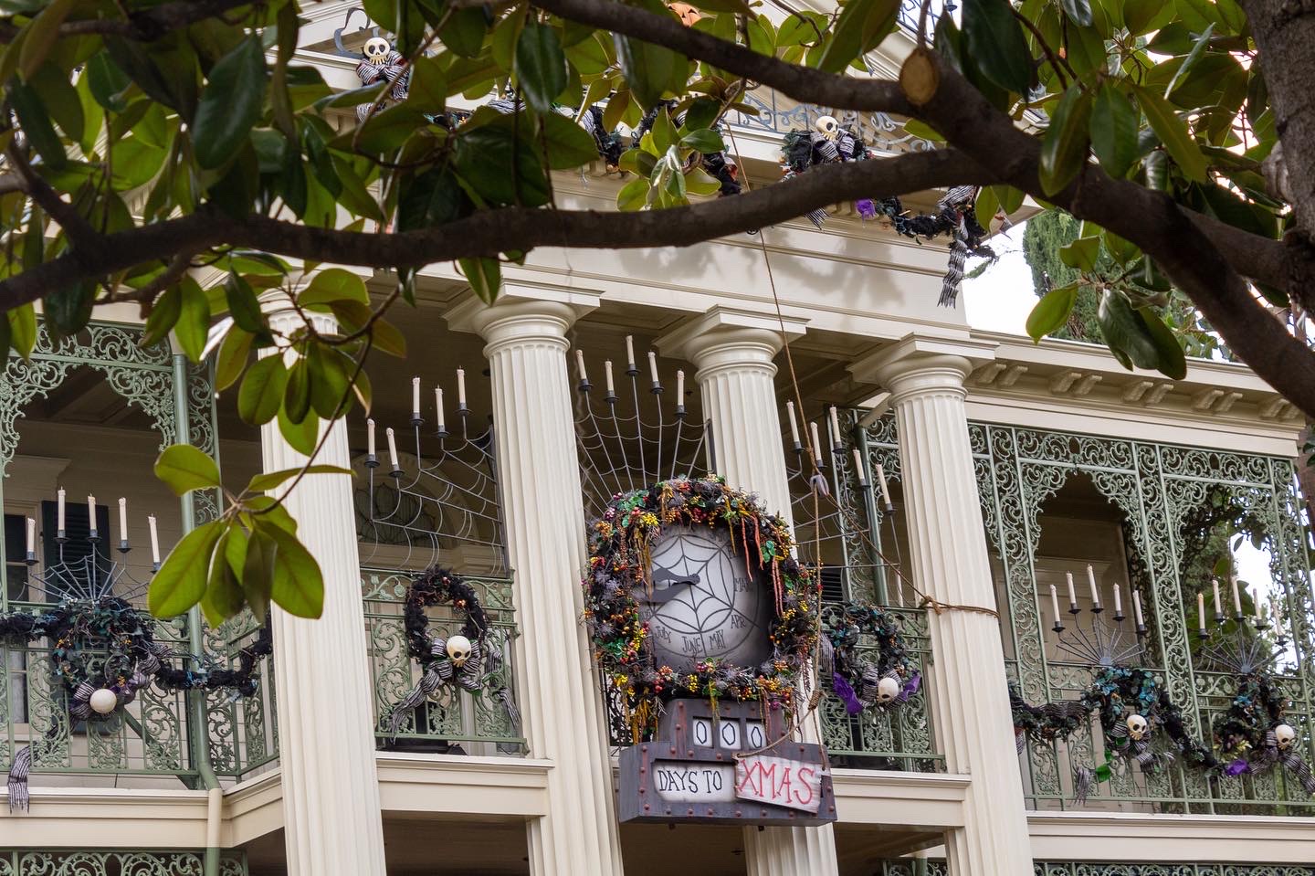 A section of the upper outside level of the Haunted Mansion, a white New Orleans-style building with pale green decorative grating, with a large holiday wreath and a sign reading O days until Xmas
