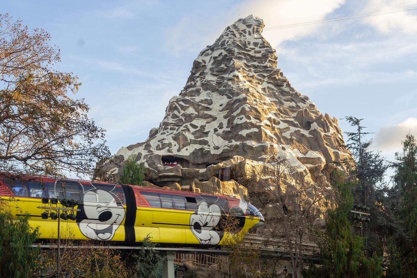 A yellow and red monorail printed with Mickey Mouse travels in front of the snowy peak of the Matterhorn Bobsleds ride under a soft blue sky