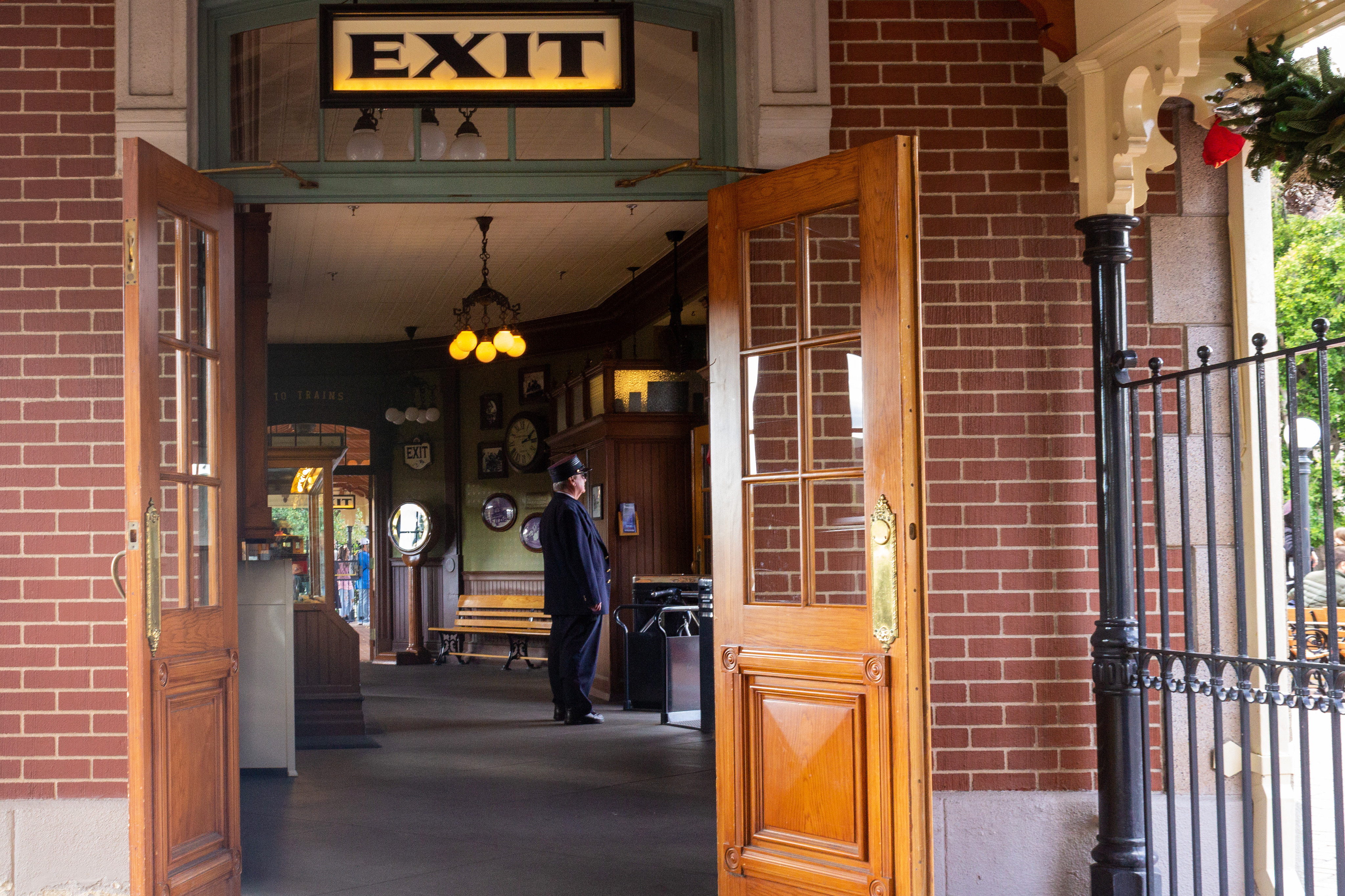 Through an open door with an exit sign, a conductor in a dark blue uniform stares ahead, waiting for a train to arrive