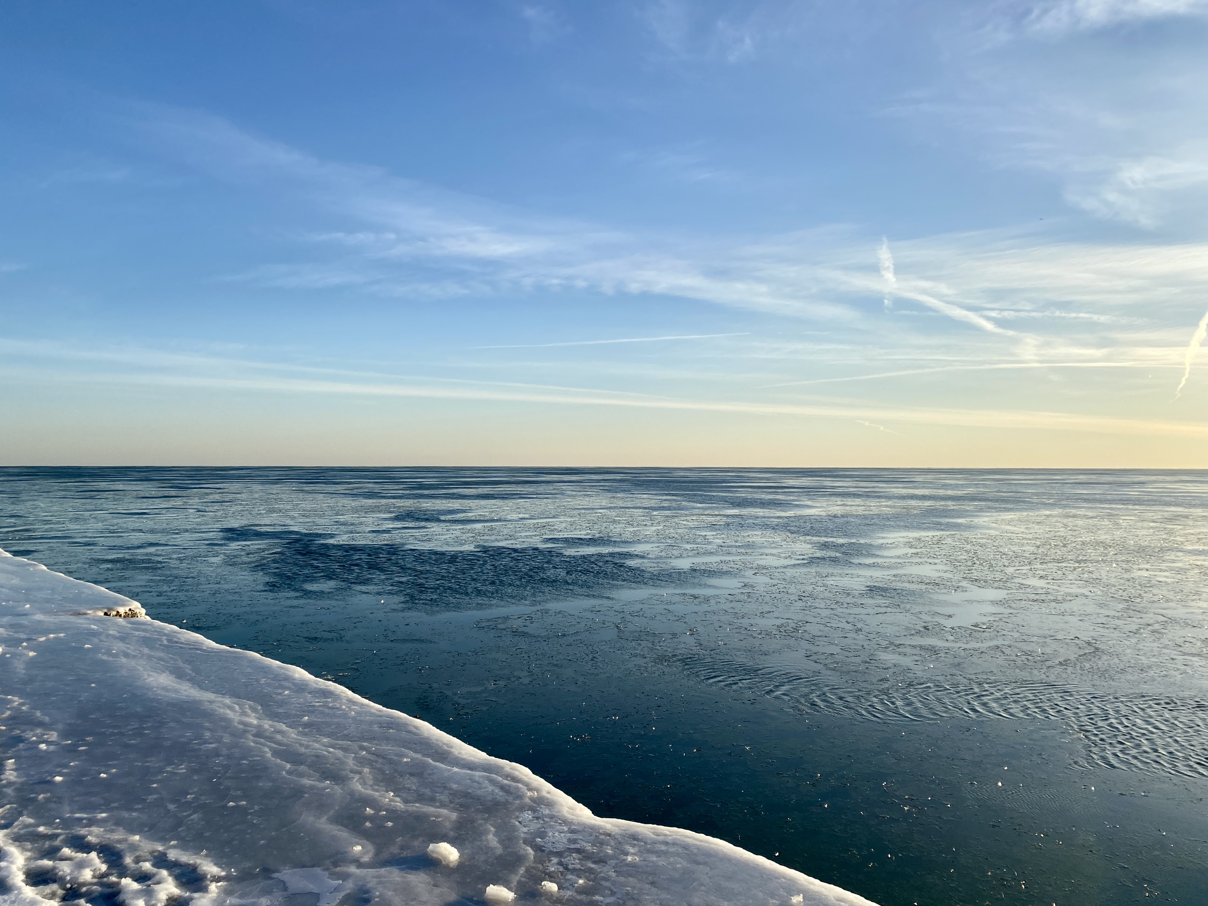 A wide lake horizon of dark blue water and blue sky with wisps of clouds and yellow with the sunrise, an bit of concrete walkway iced over at the left