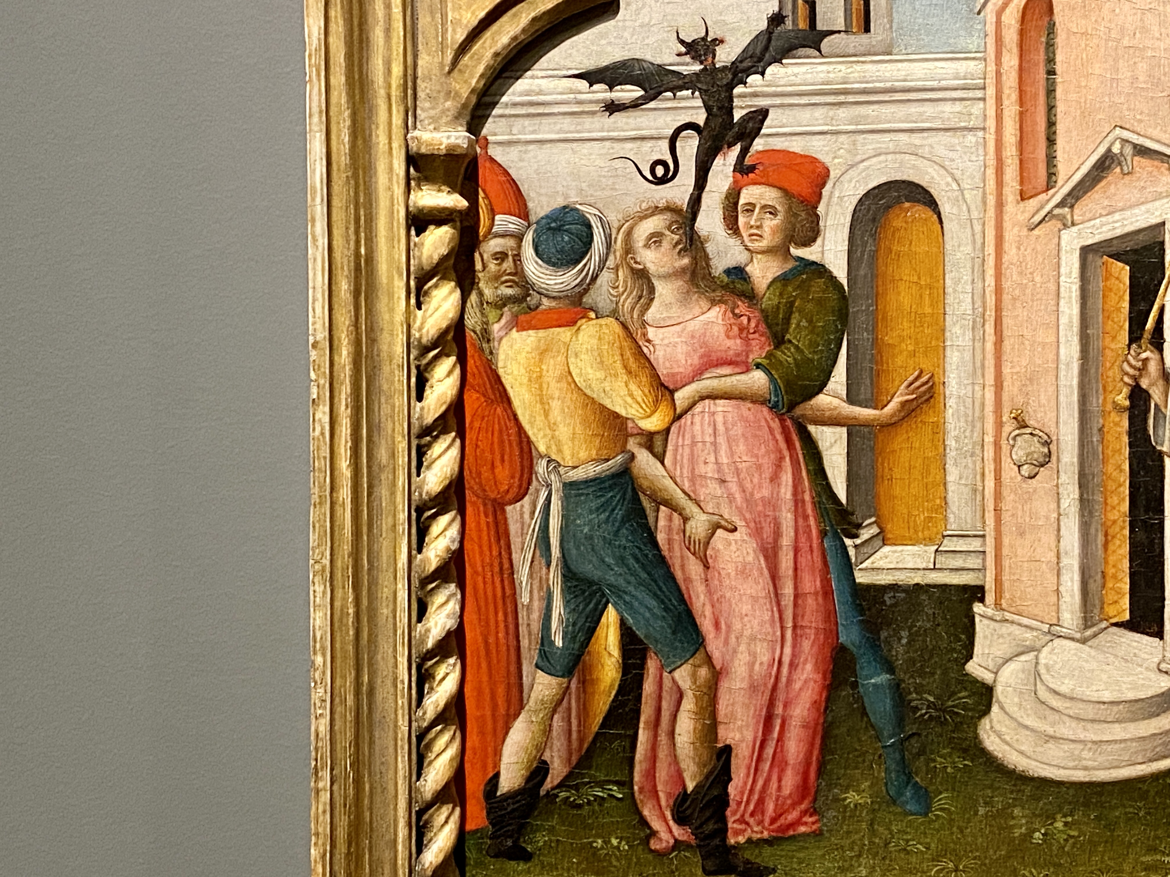 Detail of a brightly-colored medieval painting of peasants holding a young woman while a black demon floats from her mouth