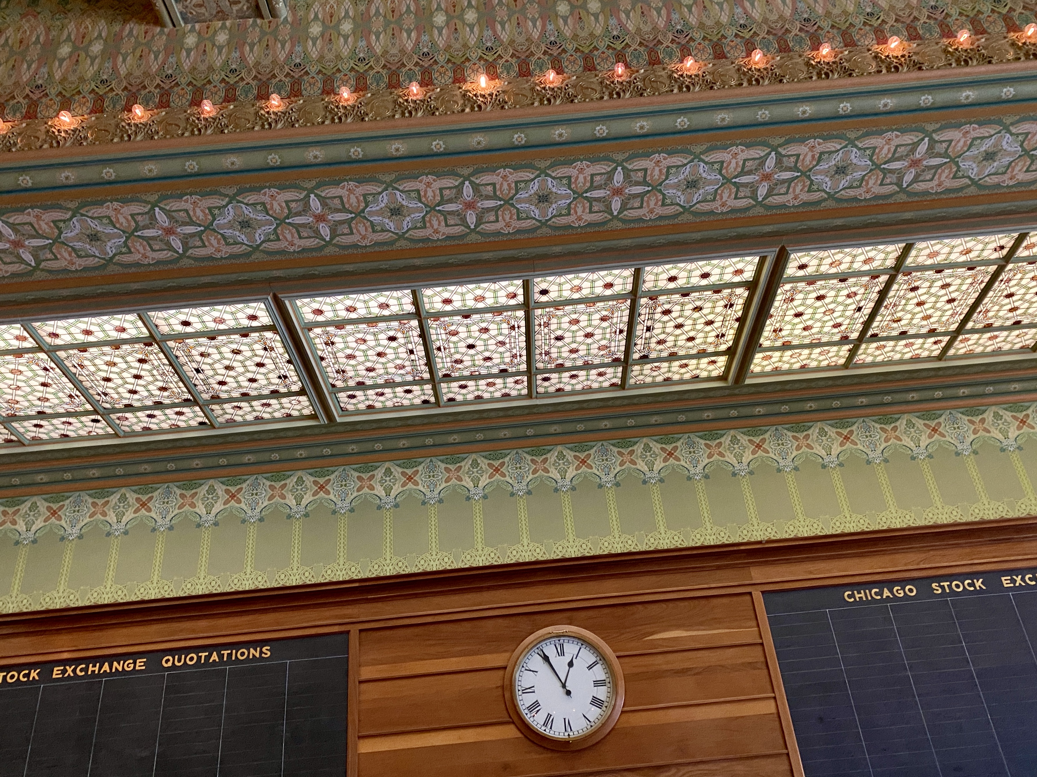 A slightly angled view of old Chicago Board of Trade stock chalkboards, wooden paneling, centered clock and ceiling intricately designed with Art Nouveau-style yellow and green flourishes