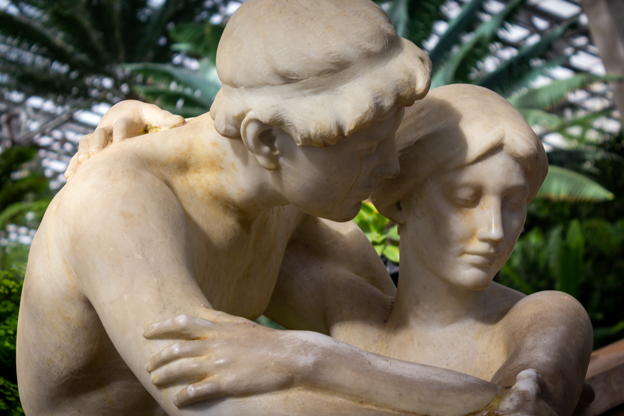 Closeup on a white marble sculpture of a young man embracing a young woman in front of greenery