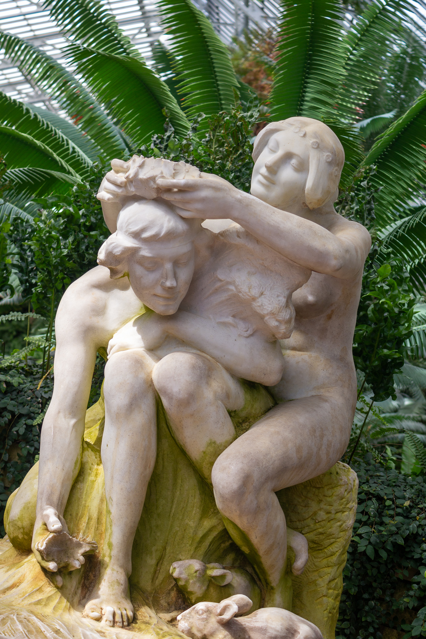 Full portrait of a white marble sculpture of two youths, one laying a wreath of leaves on the head of the other, the fronds of a palm spread above them