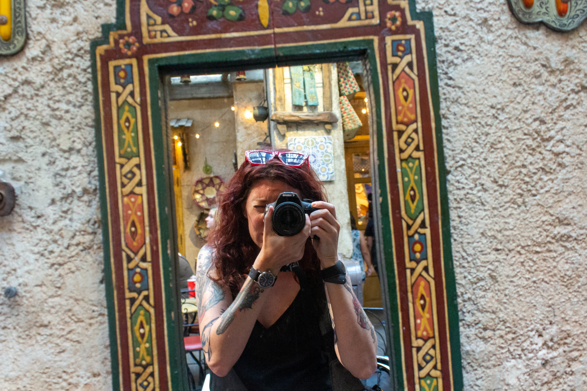 The image of a white woman with long reddish wavy hair holding a camera to her eye as captured in a tall mirror edged with colorful designs
