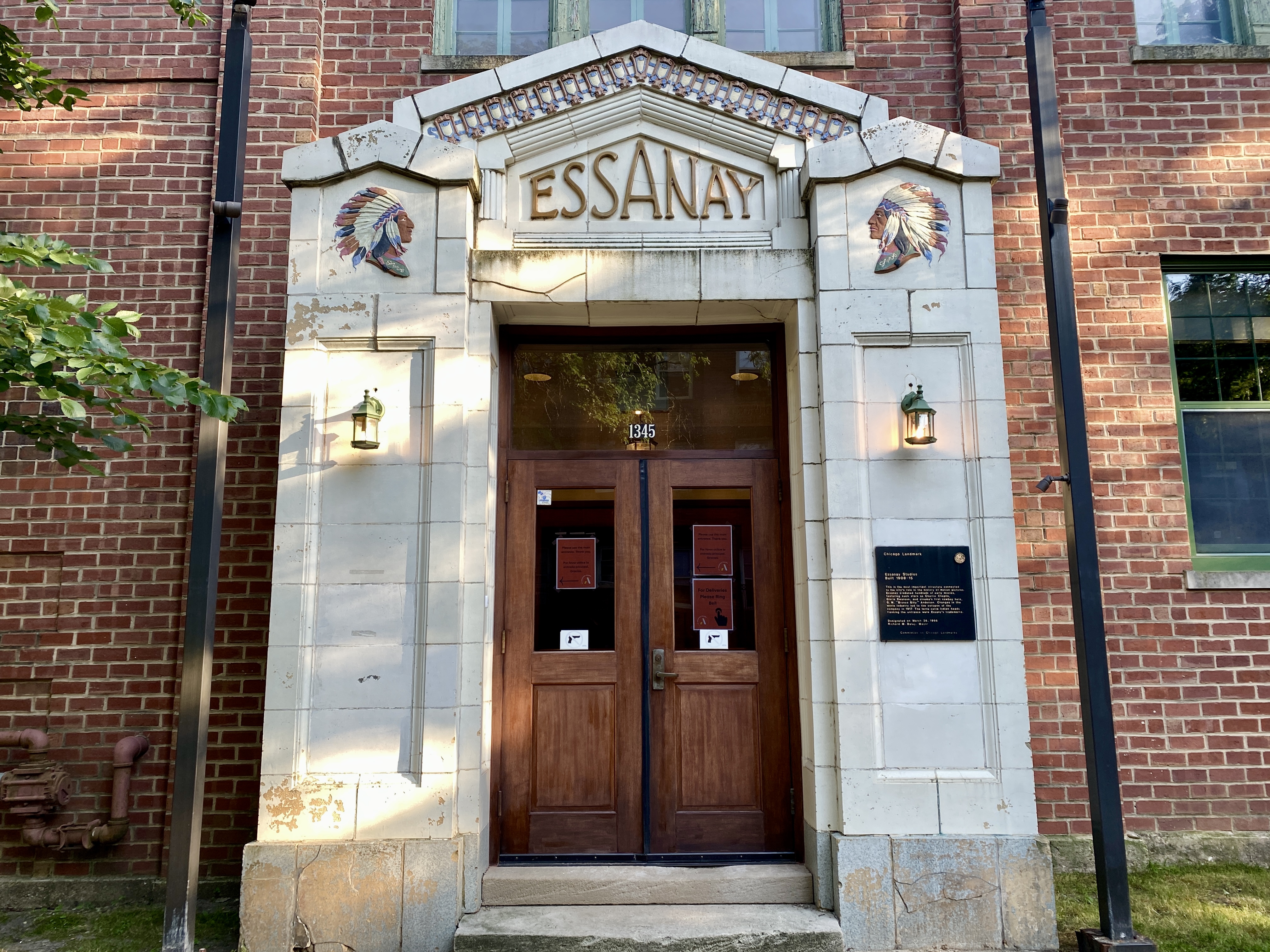 Full view an arched entryway to a brick building, matching reliefs of a Native chief on each side and over the door the stylized letters spelling ESSANAY