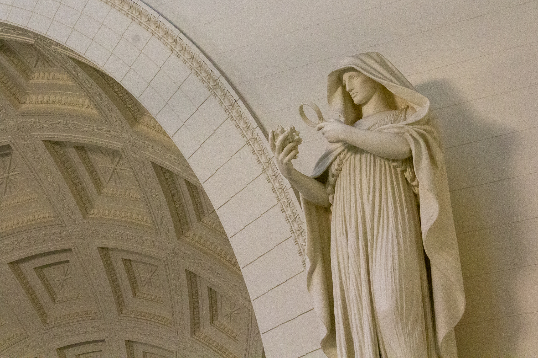 A white statue of a cloaked man examining an object in his hand, behind to the left a curve of molded, arched ceiling