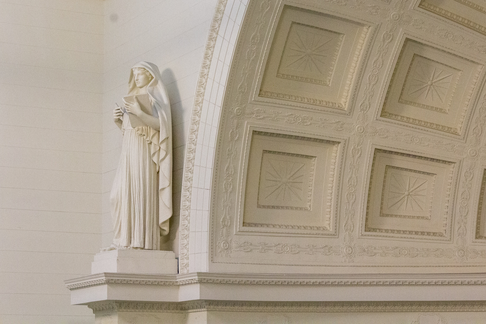 A white statue of a robed figure holding a book, to the right a curve of molded, arched ceiling