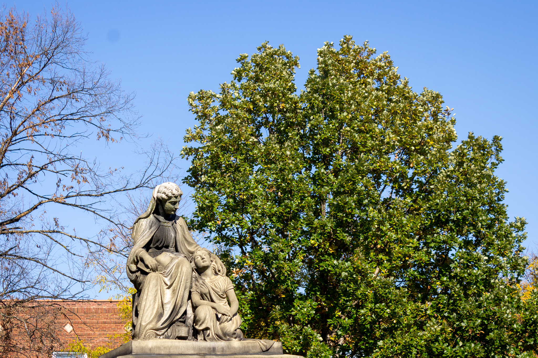 The top of a monument with a carved stone woman and child against a blue sky and large green tree