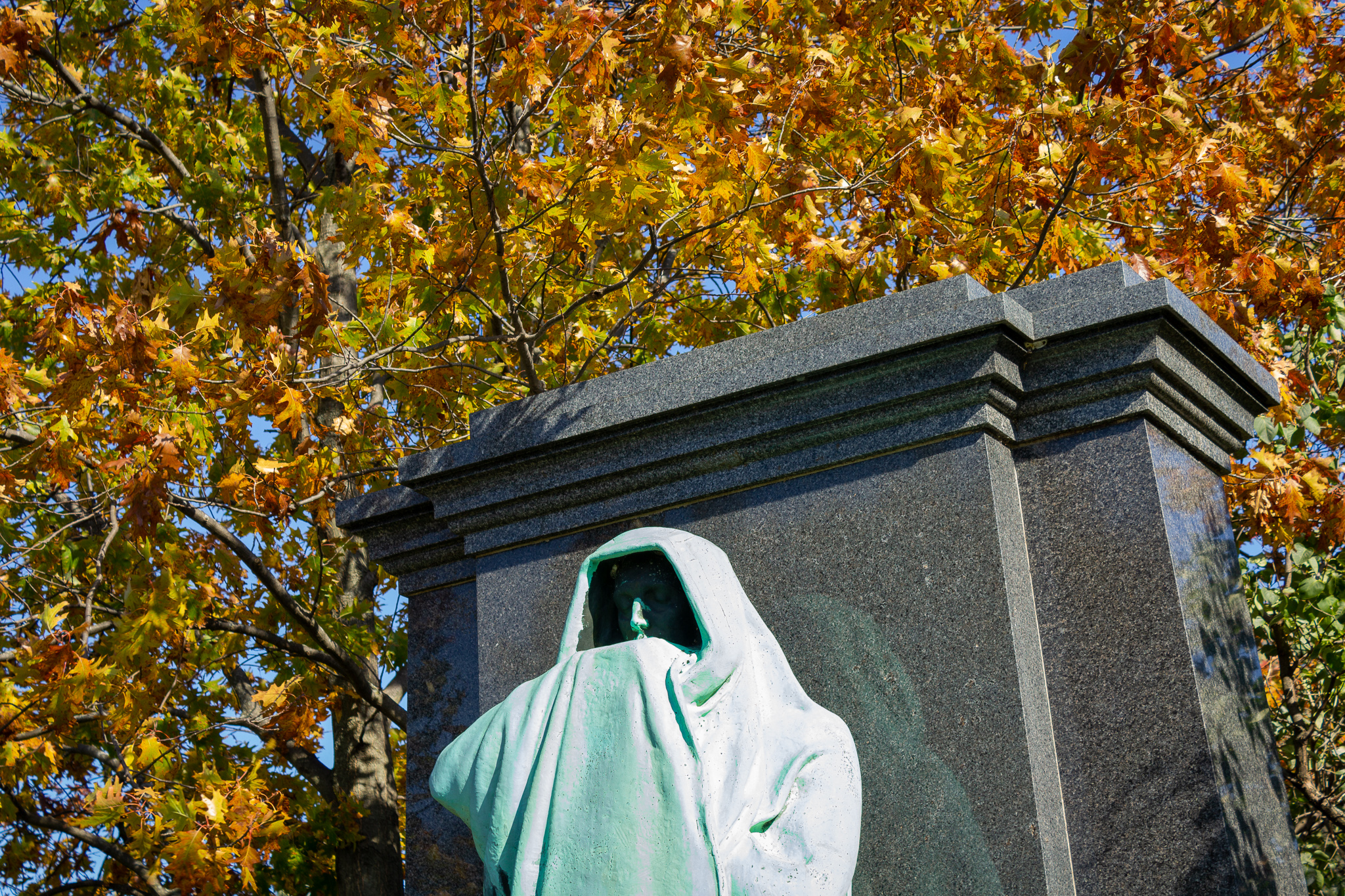 Top of a greened copper statue of a cloaked man against a polished marble monument, in the background a yellow-leafed tree