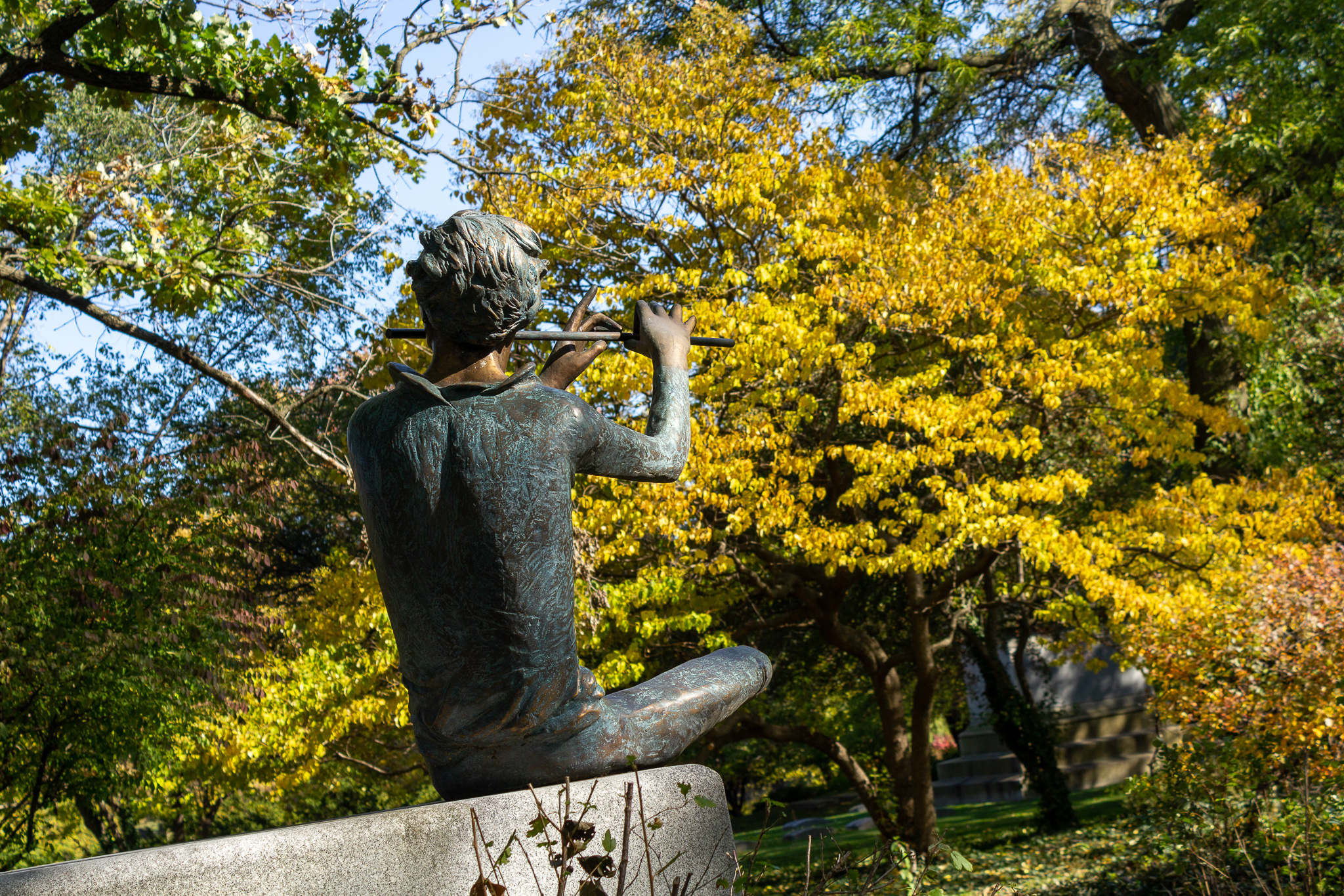 The aged bronze figure of a boy playing a flute, seated on a gravestone, yellow trees in the background