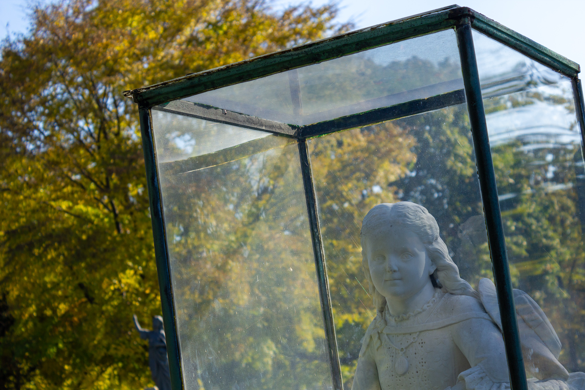 The stone figure of a young girl smiles through a glass box with yellow trees in the background