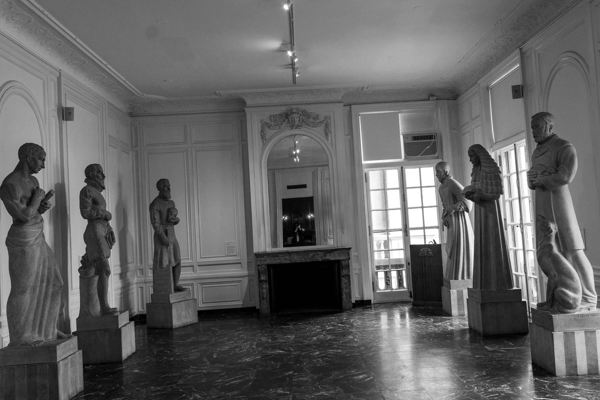 Canted black-and-white image of a large room lined with statues, a black marble floor and in the center a fireplace topped with a mirror, in which the distant image of the photographer is reflected