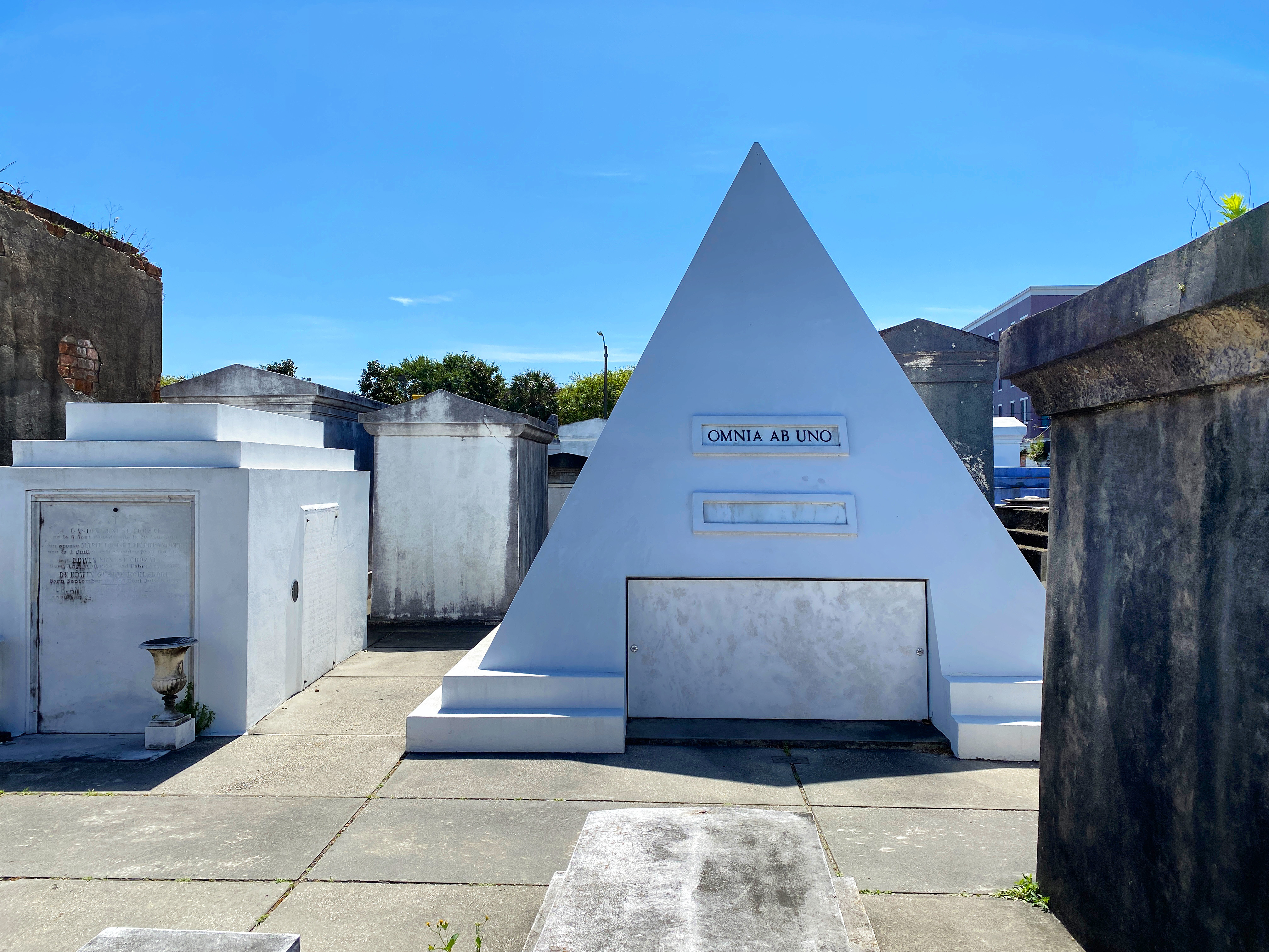 A white, pyramid-shaped tomb in among other tombs in a cemetery, inscription on it reads: “Omnia ab uno”
