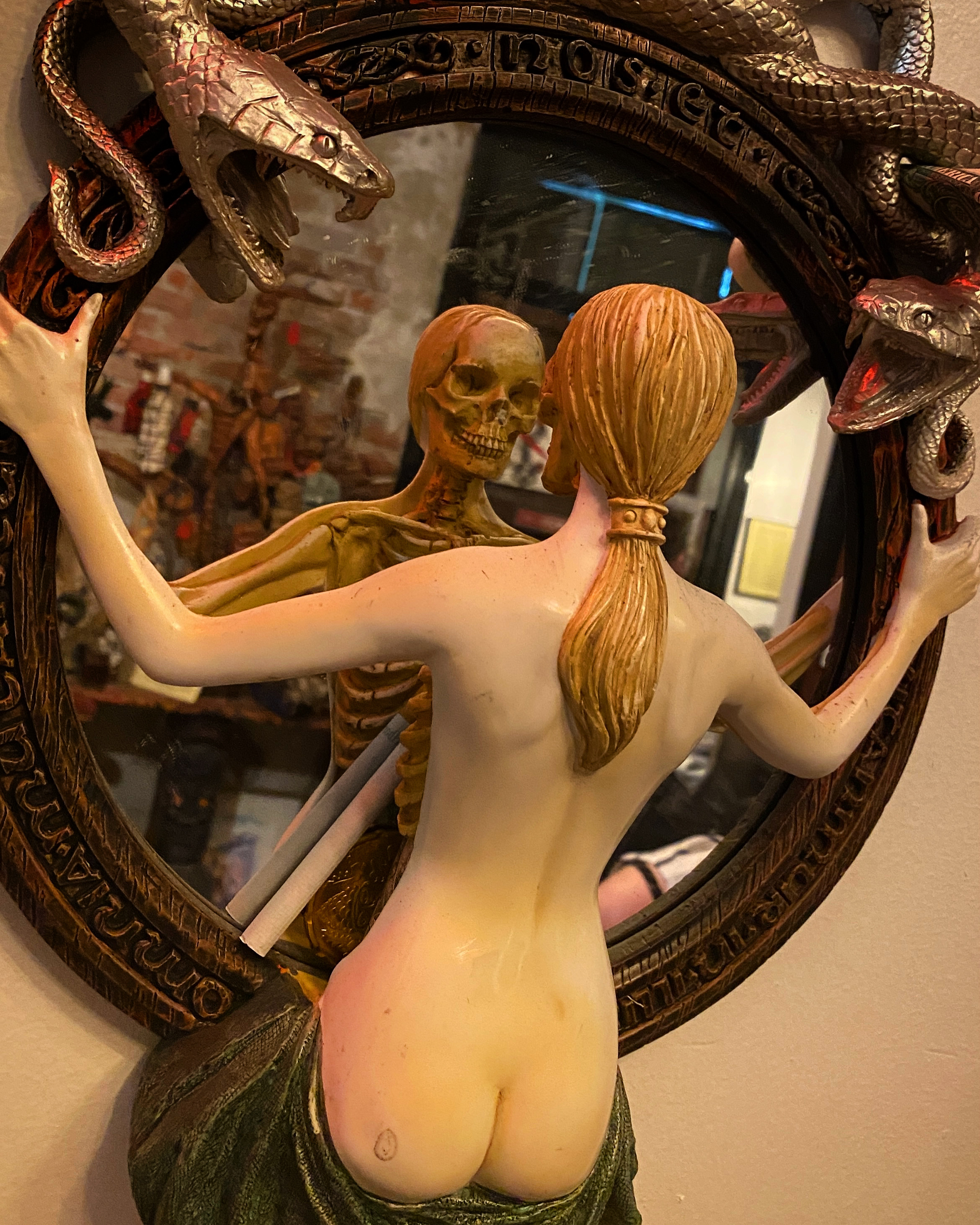 A round mirror topped with molded snakes, the figure of a naked white woman sculpted to look into the mirror, but her reflection shows a skeleton 