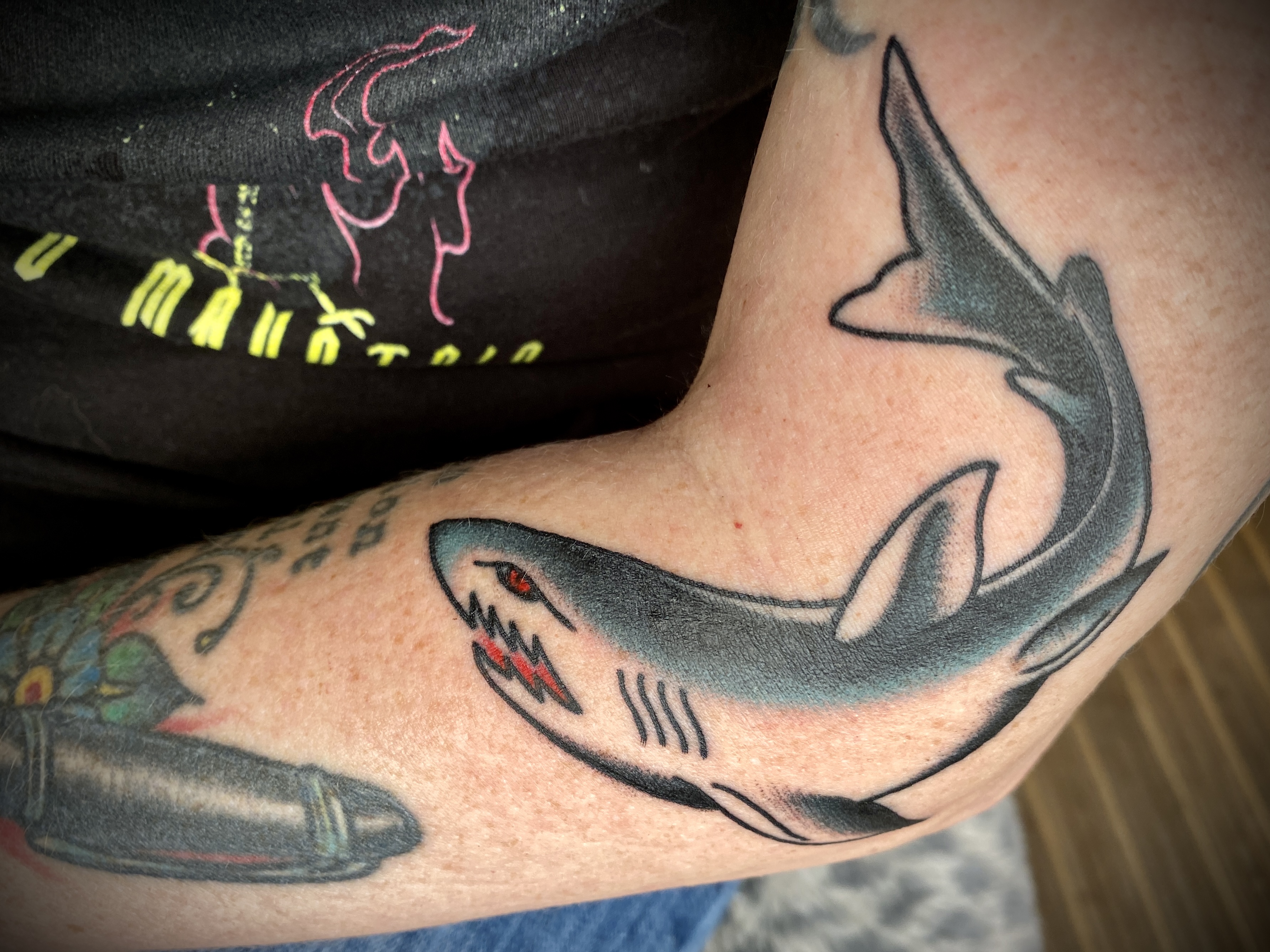 A white woman's bent elbow with a curved shark tattoo, shaded in dark grey and blue with a red eye and red jagged mouth
